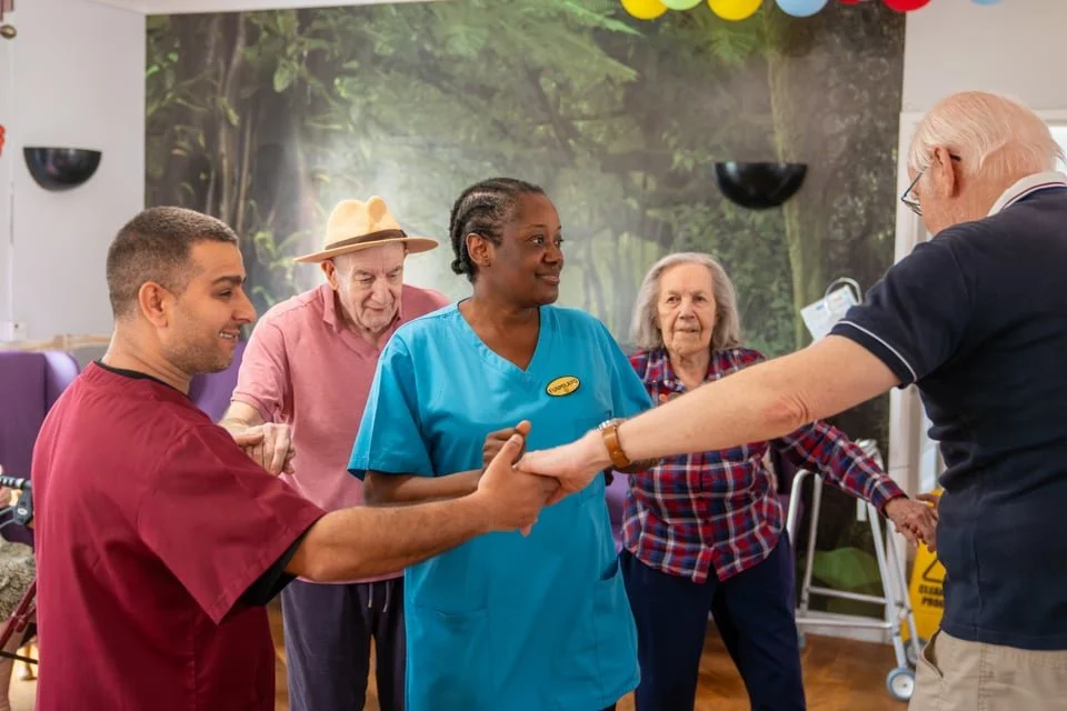 A group of five people in a care facility, including a nurse in blue scrubs and four elderly residents, are standing in a cheerful circle. They are shaking hands and smiling, with a nature-themed mural and colorful balloons in the background.