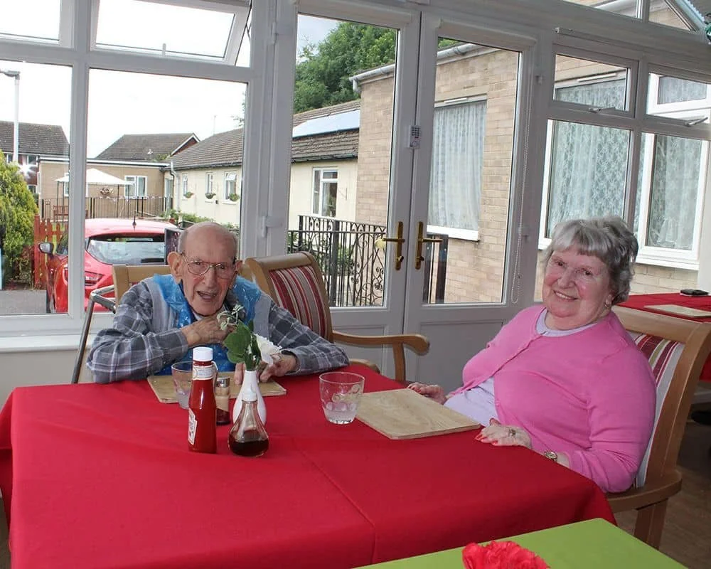 An elderly man and woman sitting at a red-covered dining table inside a bright sunroom, smiling. The man is eating, wearing glasses and a checkered shirt, while the woman is wearing a pink sweater. There are condiments, a glass of water, and a flower in a vase on the table. Outside the sunroom are houses and greenery.
