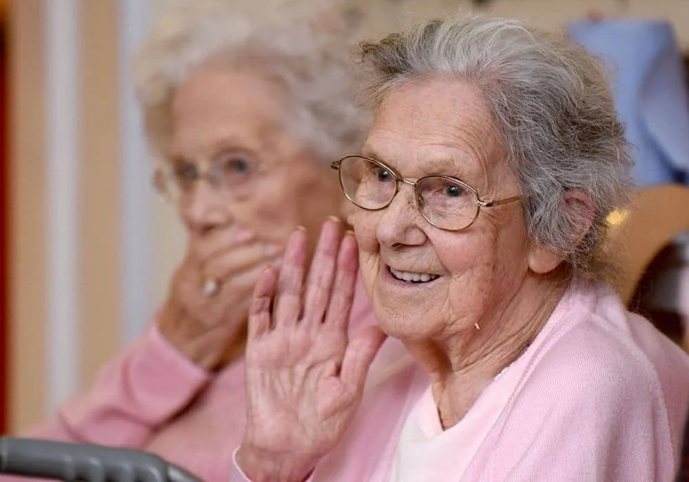 Two elderly women sitting closely, one smiling and waving, the other with hand near mouth, both wearing pink clothing in a cozy environment.