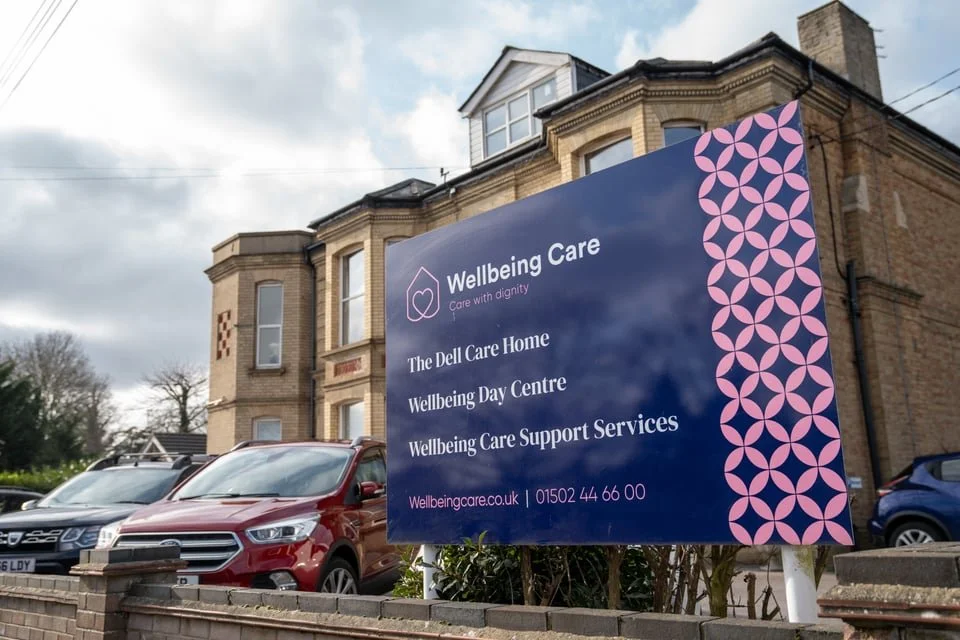 A large blue sign for Wellbeing Care outside a brick building with cars parked nearby. The sign lists services including The Dell Care Home, Wellbeing Day Centre, and Wellbeing Care Support Services, with a pink geometric border on the right side.