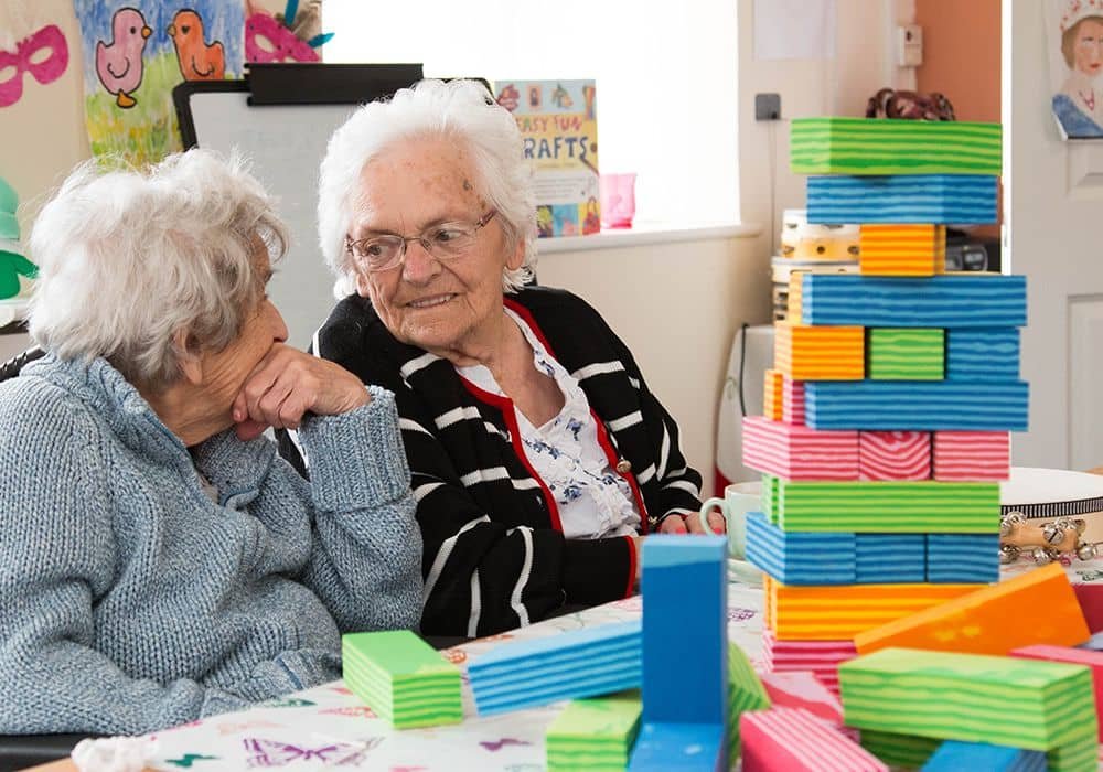 Two elderly women sitting at a table with a large stack of colorful wooden blocks, engaged in conversation.