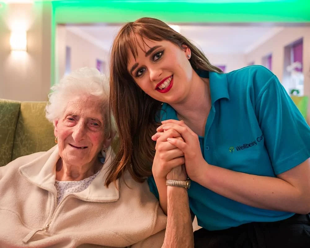 A young woman and an elderly woman smiling, holding hands, and sitting on a sofa in a well-lit room.