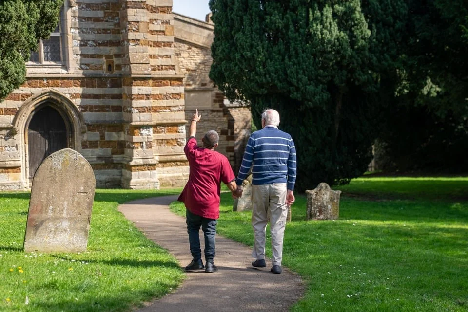 An elderly man and a middle-aged man walk hand in hand along a path outside a church, with the younger man pointing upward.