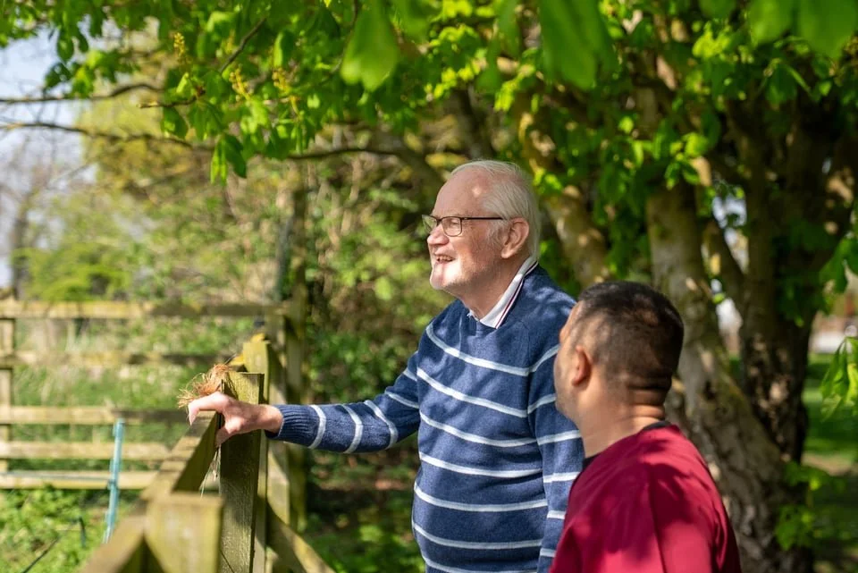 An elderly man and a young man sharing a joyful moment outdoors beneath a leafy tree, with a wooden fence in the background.