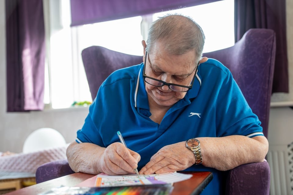 An elderly man with glasses and a watch sitting in a purple armchair, writing or drawing on paper with a pen at a table in a sunlit room.