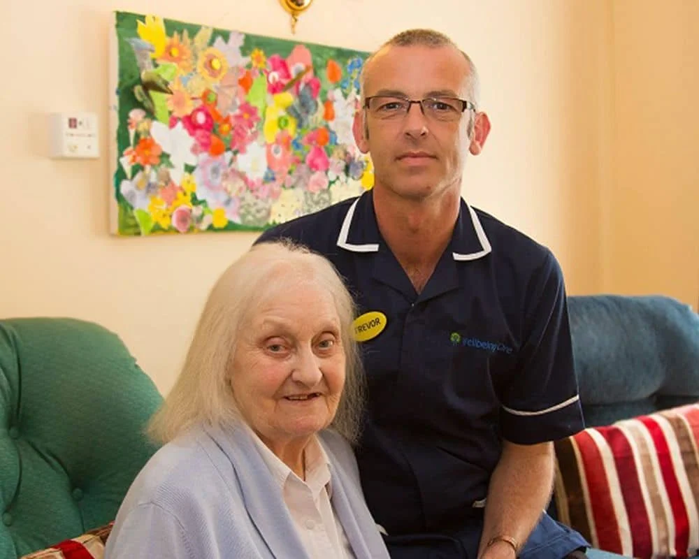 An elderly woman with white hair sitting on a green couch, with a man standing beside her. The man is wearing glasses, a navy blue polo shirt with a name tag that says 'Trevor' and the logo of 'realcare.' The background features a colorful floral painting on the wall.