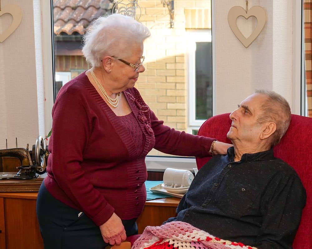 An elderly woman dressed in a maroon sweater and pearl necklace is gently looking at an elderly man sitting in a red armchair. The woman has her hand on the man's shoulder, and they are looking into each other's eyes inside a cozy room with heart-shaped decorations on the wall behind them.