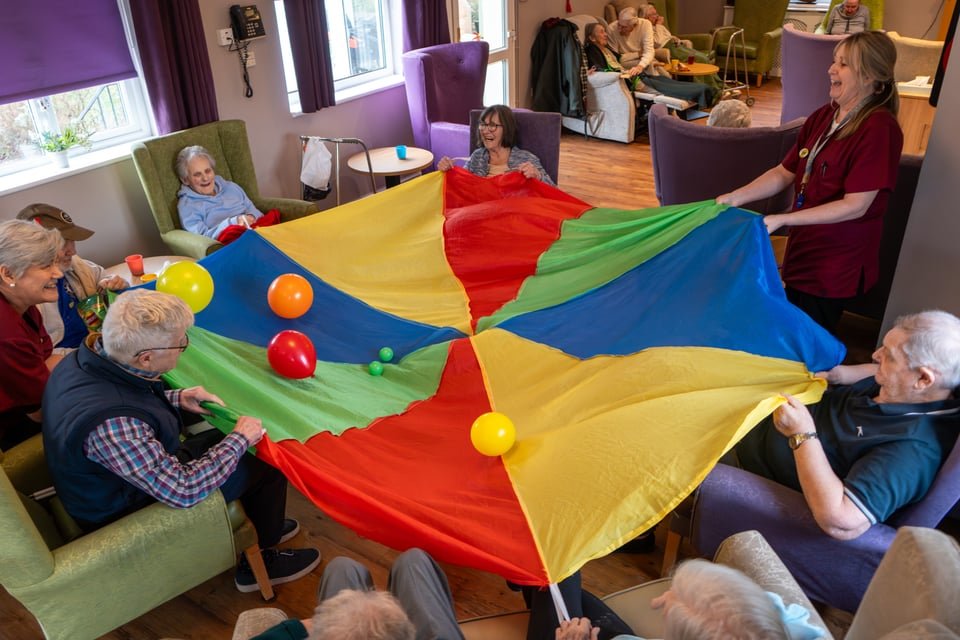 Elderly residents and caregivers playing with a colorful parachute with balls in a communal space of a care home.