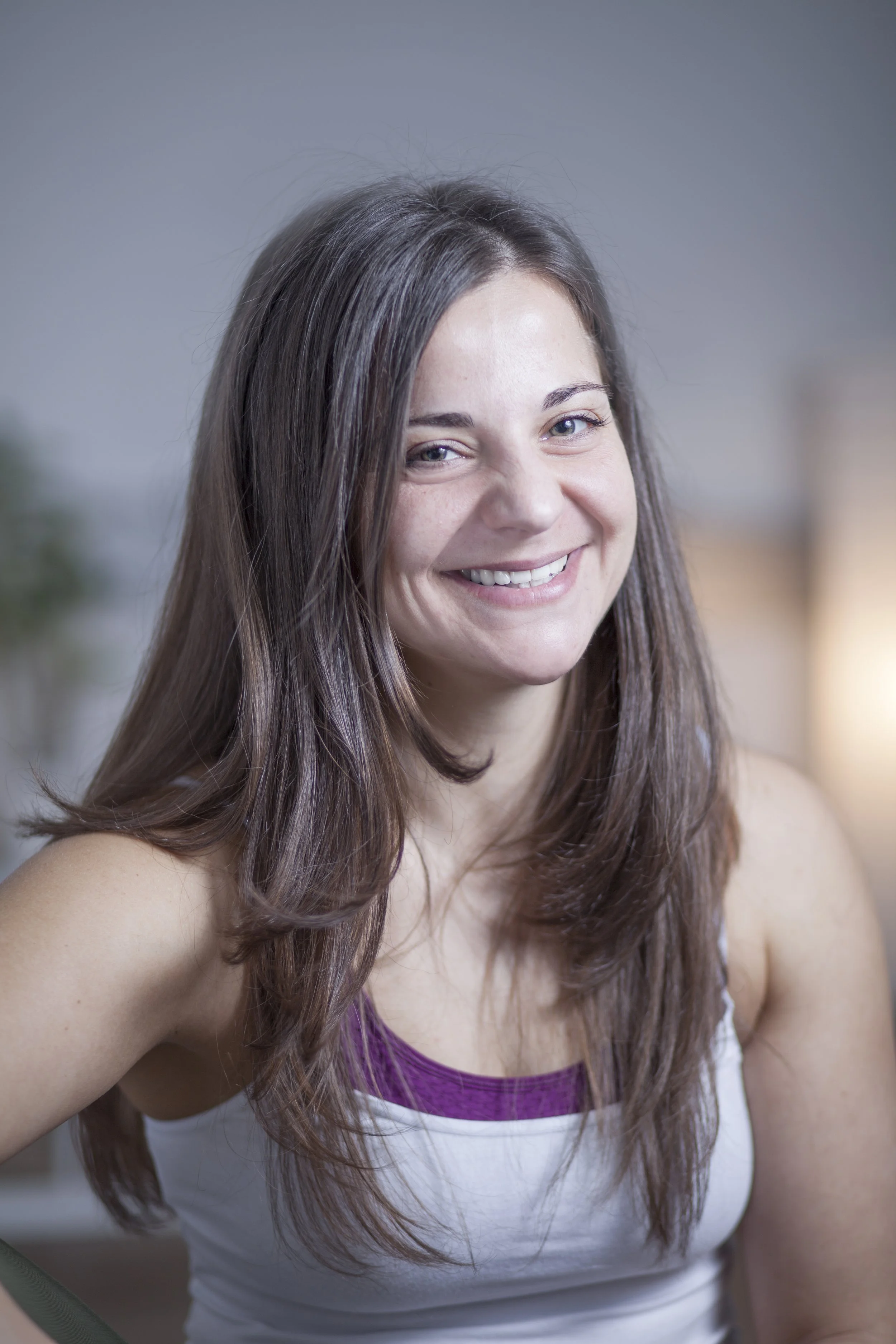 Nahaufnahme einer lachenden jungen Frau mit langem braunem Haar, trägt ein weißes Top mit lila Akzent, steht vor einem unscharfen Hintergrund.
