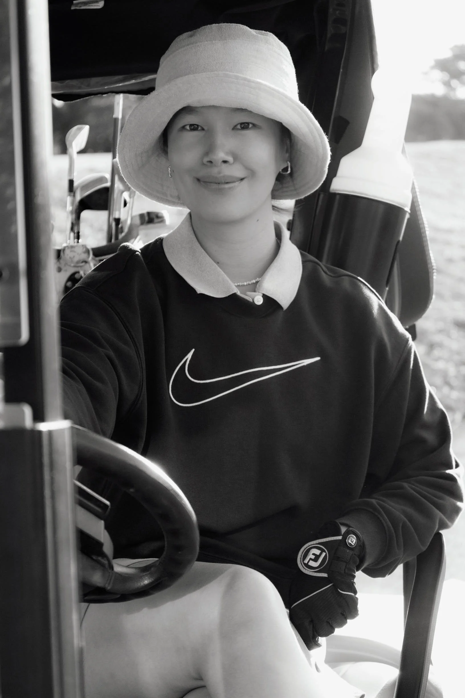 A woman sitting in a golf cart, wearing a wide-brimmed hat, a Nike sweatshirt, and golf gloves, smiling at the camera.