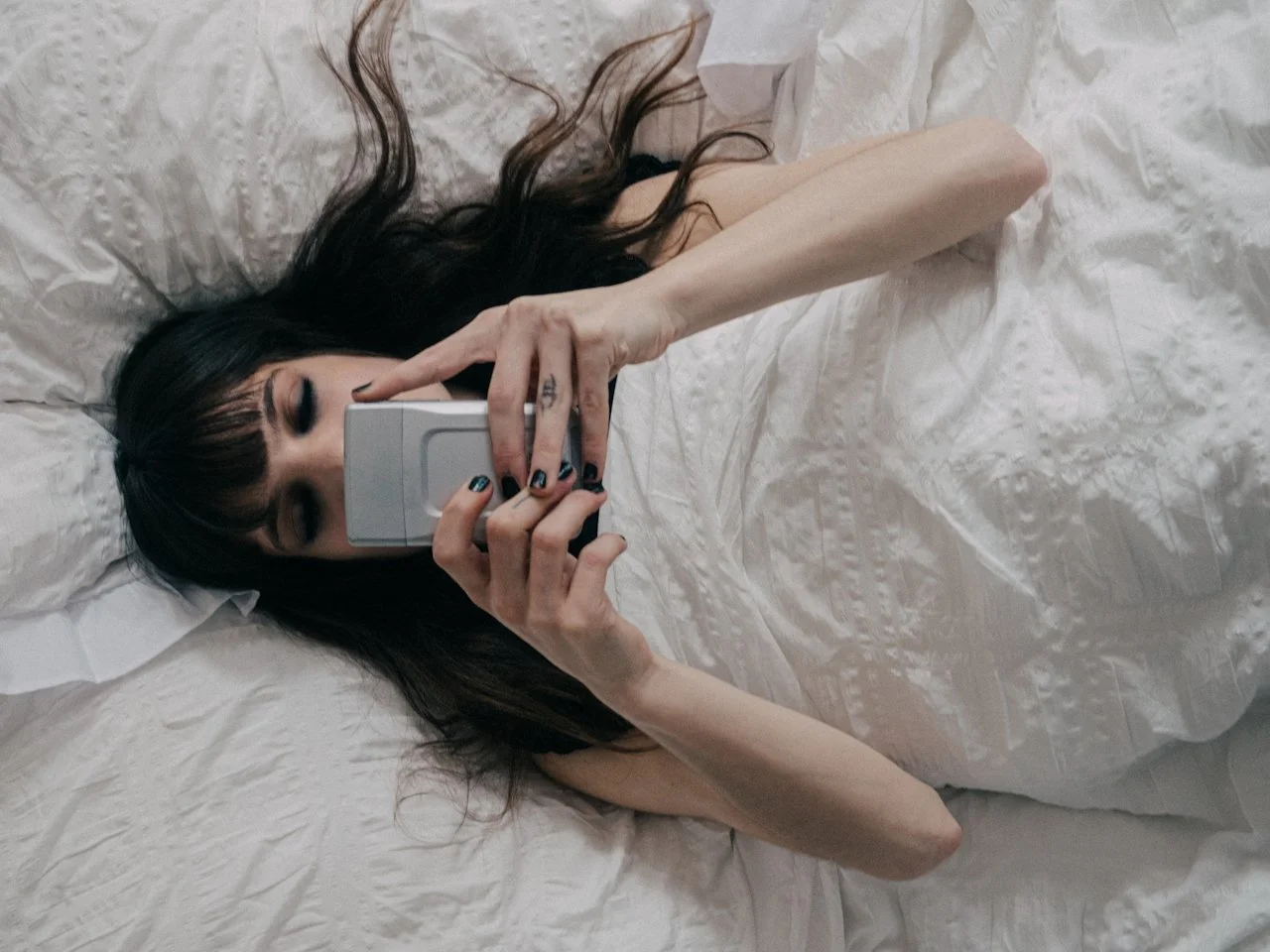 A woman with dark hair and makeup lying on a bed, holding a flip phone in front of her face to take a selfie.