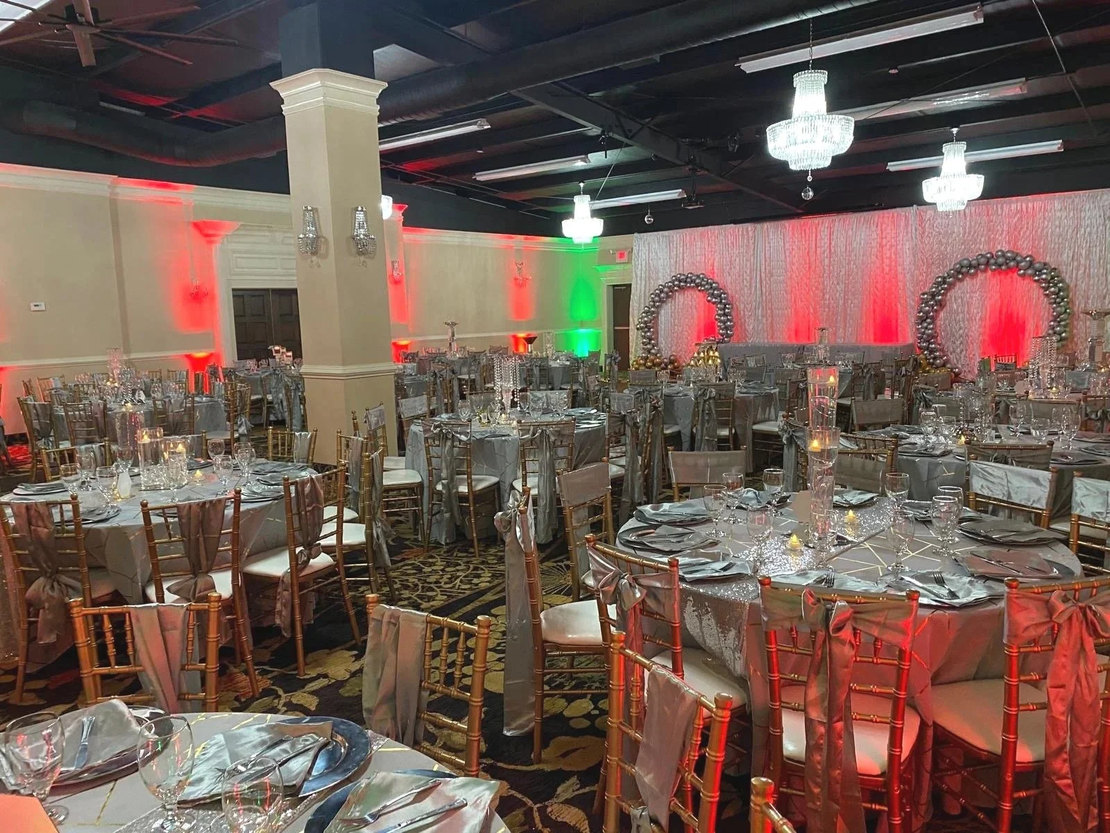 A banquet hall decorated for a celebration with round tables set with glassware, napkins, and candles. The room has elegant chandeliers, balloon arches at the back, and colorful lighting with red and green accents.