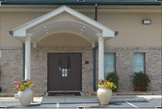 Entrance of a building with a covered porch, two potted plants with colorful flowers on each side of the door, and two windows with bushes in front of them.