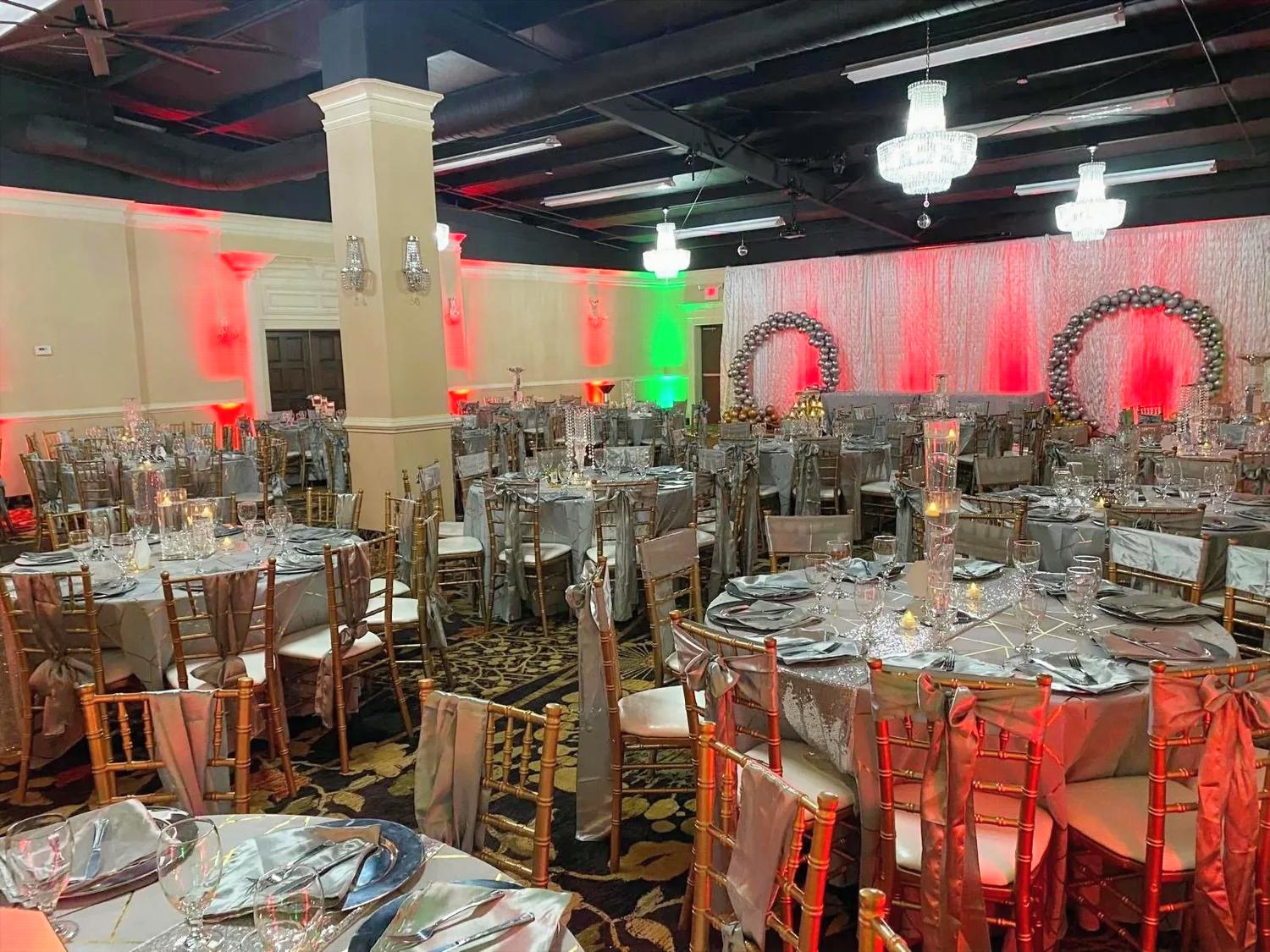 Elegant banquet hall decorated with tables set for a party or wedding reception, featuring gold chairs, centerpieces, and a backdrop with Christmas wreaths and red and green lighting.