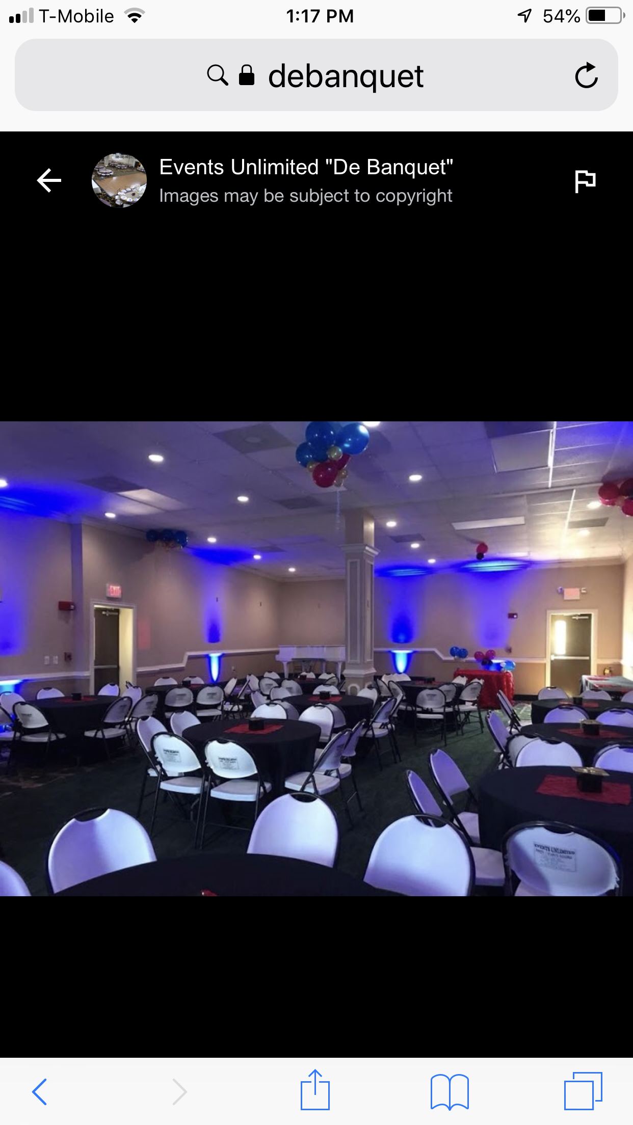 A decorated banquet hall with round tables covered with black tablecloths, white chairs, and blue and red balloon decorations. The room has blue uplighting on the walls, a white piano in the corner, and balloons hanging from the ceiling.