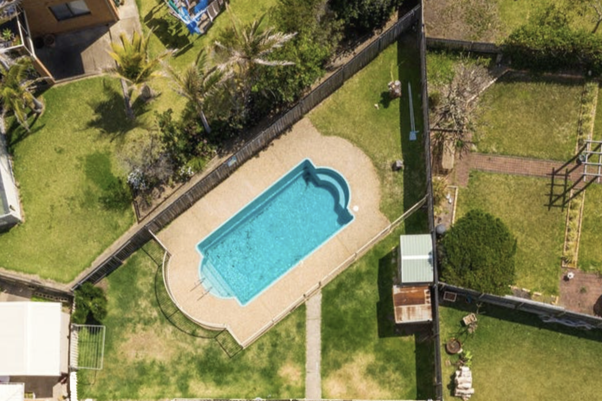 Aerial view of a backyard with a swimming pool, surrounded by a fenced grassy area, trees, and garden beds.