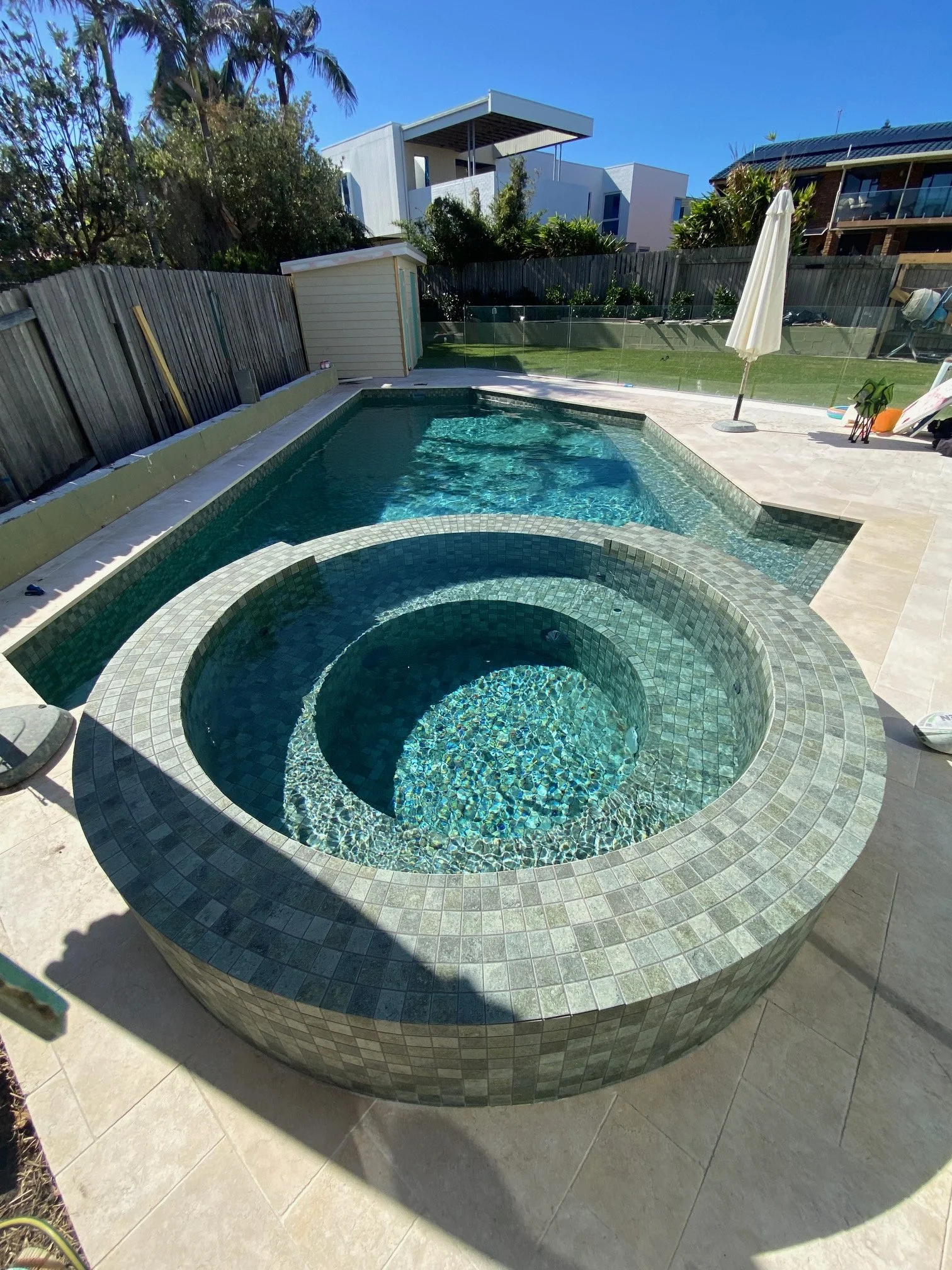 Backyard swimming pool with a spa, surrounded by a paved deck and a wooden fence. There is a white umbrella and yard equipment on the right side, with trees and modern houses in the background under a clear blue sky.