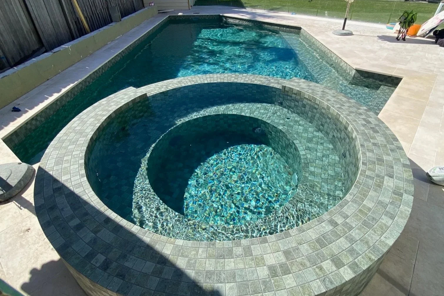 Custom concrete swimming pool with a built-in spa, surrounded by paved area in a backyard, with a fence, lawn, and some backyard items in the background.