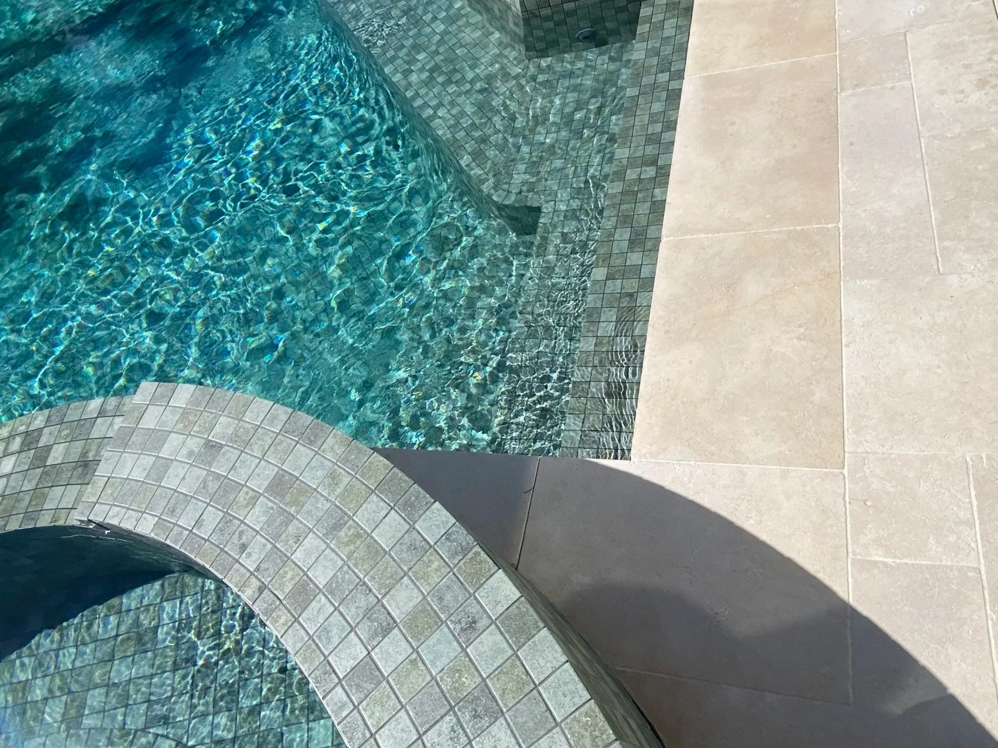 A close-up view of a swimming pool's corner with torquoise tile lining, showing the water's surface and the limestone paved surround
