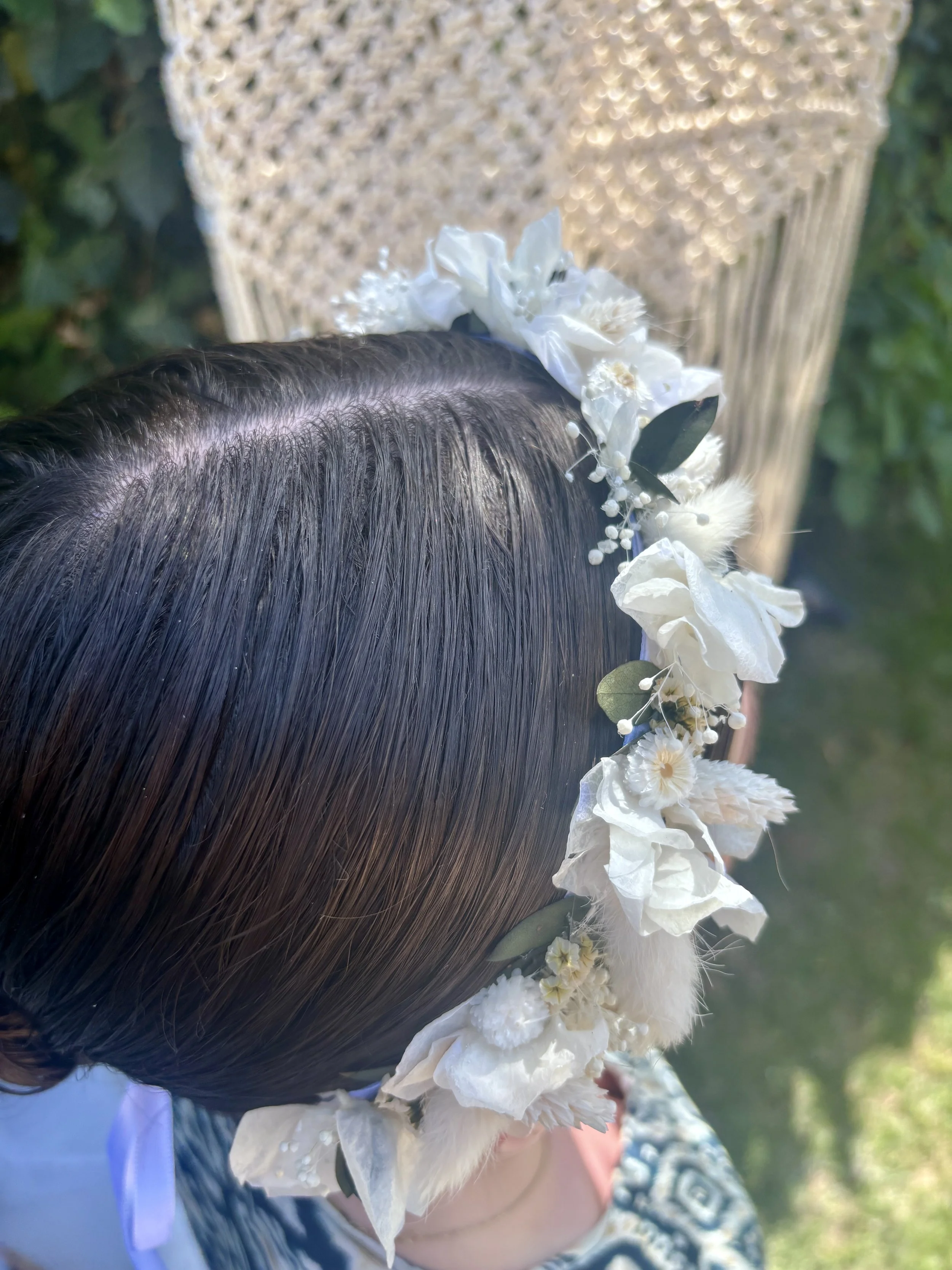 Fille avec une couronne de fleurs blanches autour de la tête, vue de profil, portant une robe à motifs, dans un environnement extérieur en plein jour.