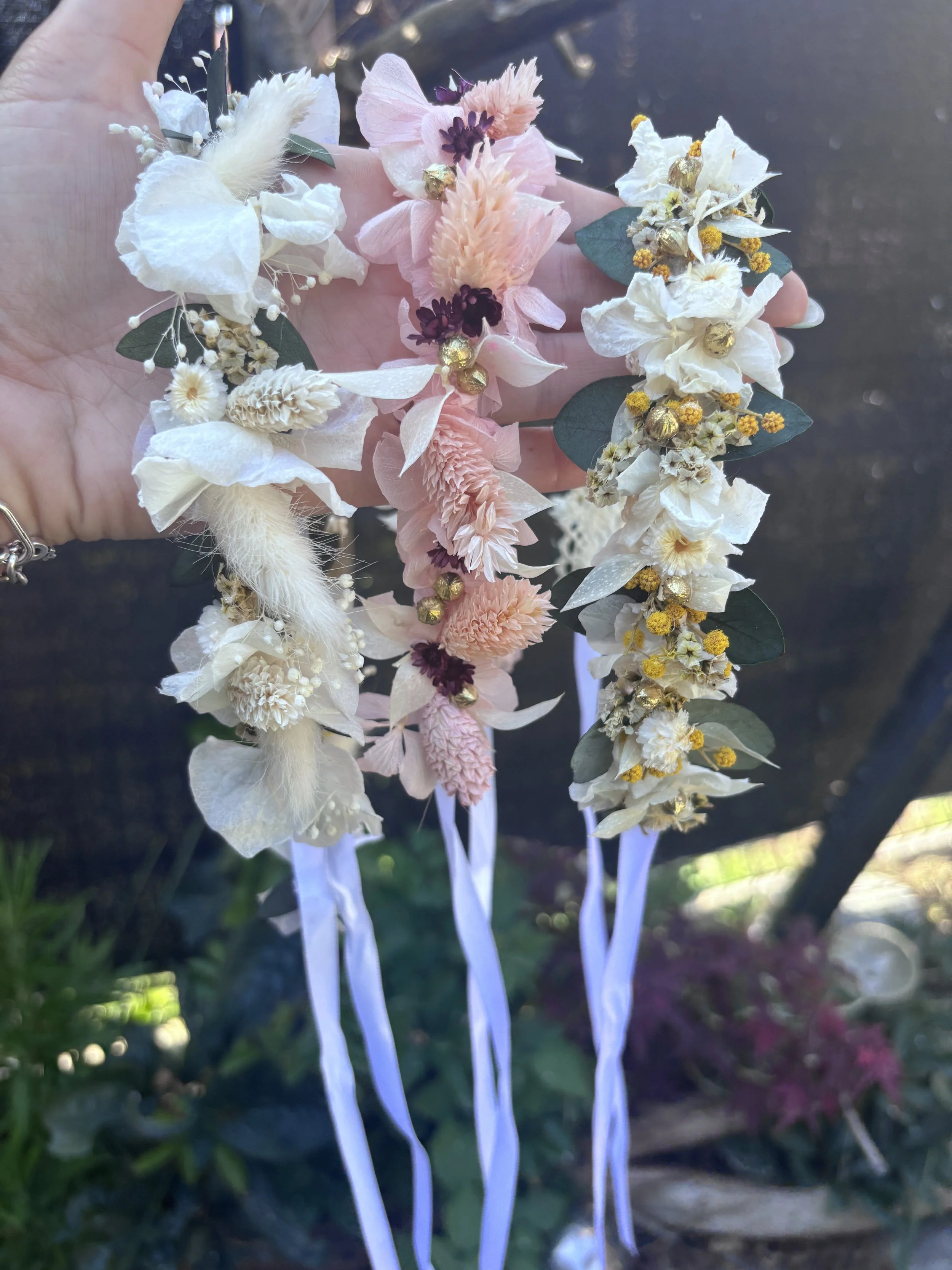Une personne tient une couronne de fleurs séchées composée de fleurs blanches, roses et jaunes avec des feuilles vertes et des rubans blancs en dessous.