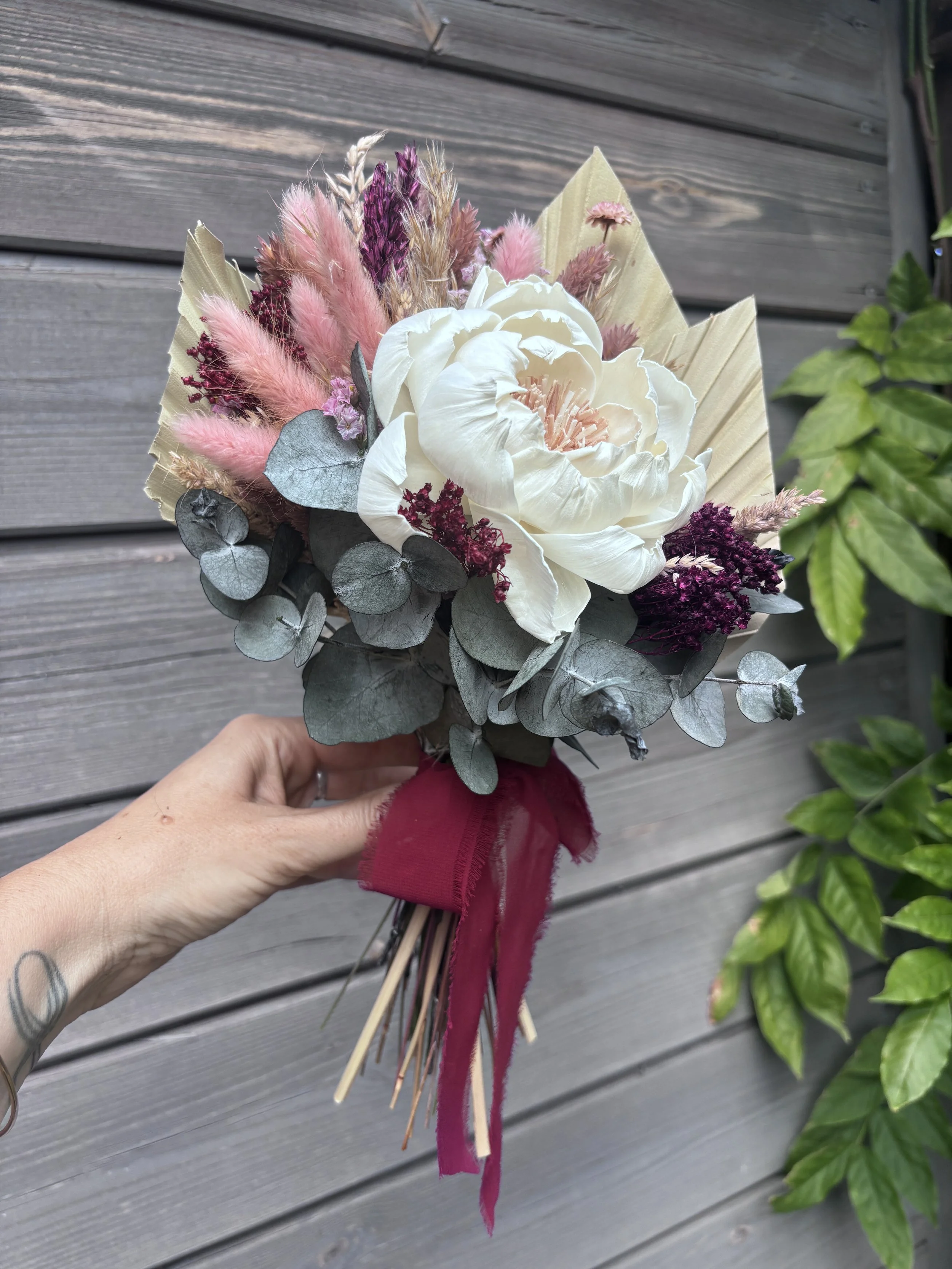 Bouquet de mariée en fleurs séchées et stabilisées, composé d’une grande pivoine ivoire en bois de Sola, d’eucalyptus, de lagurus rose poudré, de fleurs bordeaux et de palmes naturelles. Un bouquet romantique et durable, idéal pour un mariage bohème 