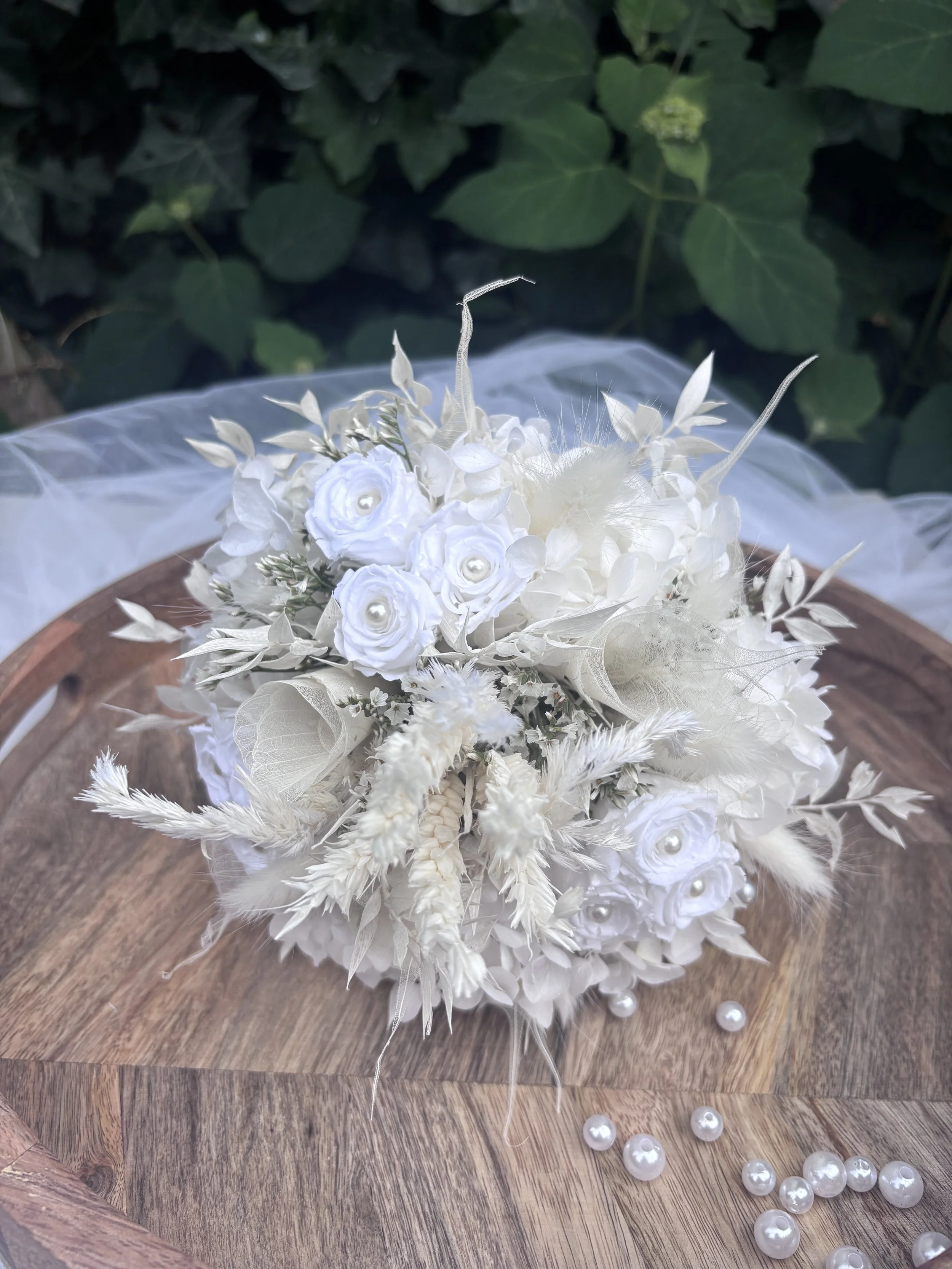 Bouquet de mariée en fleurs séchées et stabilisées blanches, réalisé artisanalement dans un esprit bohème chic et romantique. Cette création florale associe hortensias stabilisés, roses éternelles ornées de perles, arums délicats, feuillages blanchis