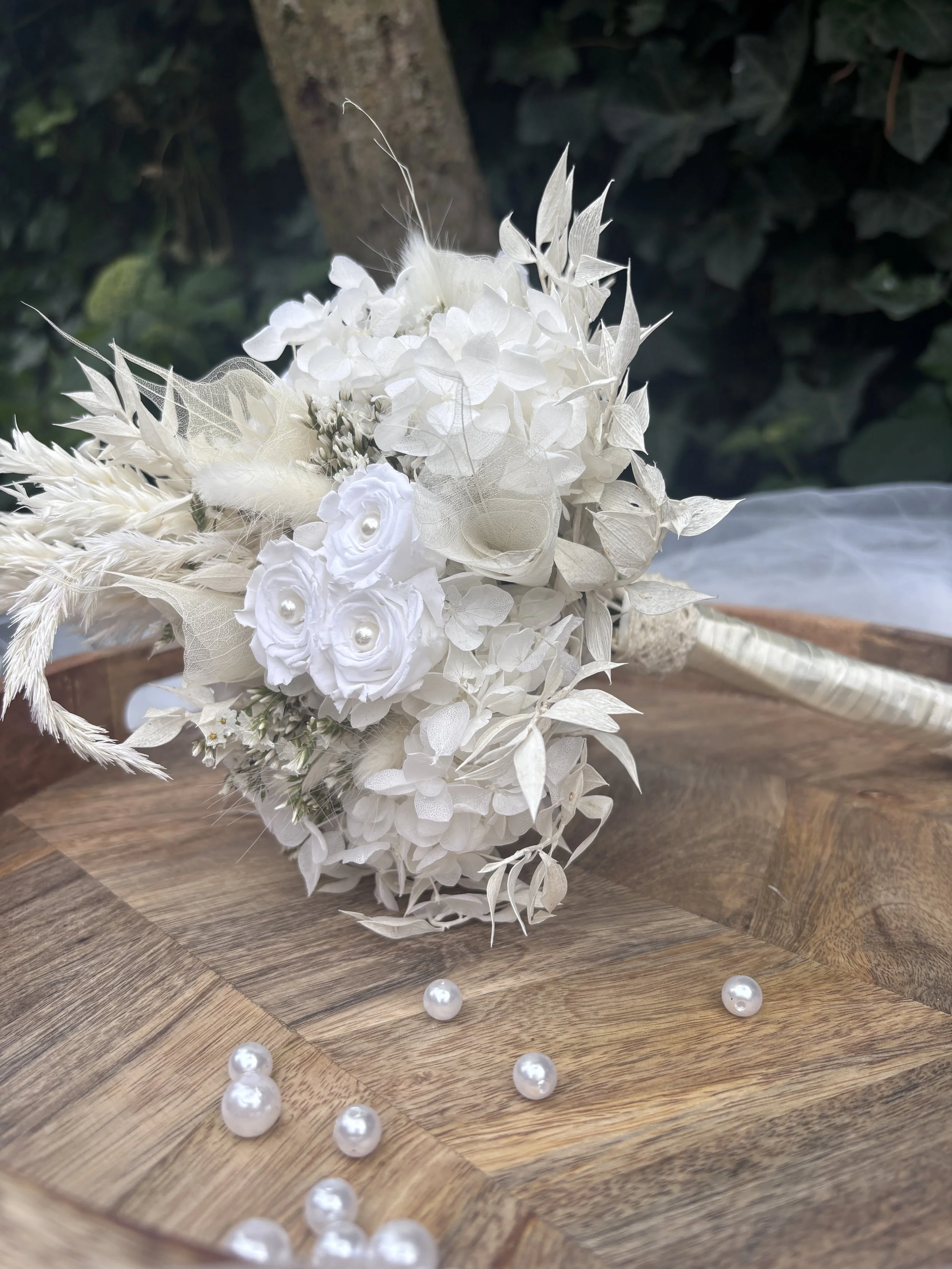 Bouquet de mariée en fleurs séchées et stabilisées blanches, réalisé artisanalement dans un esprit bohème chic et romantique. Cette création florale associe hortensias stabilisés, roses éternelles ornées de perles, arums délicats, feuillages blanchis