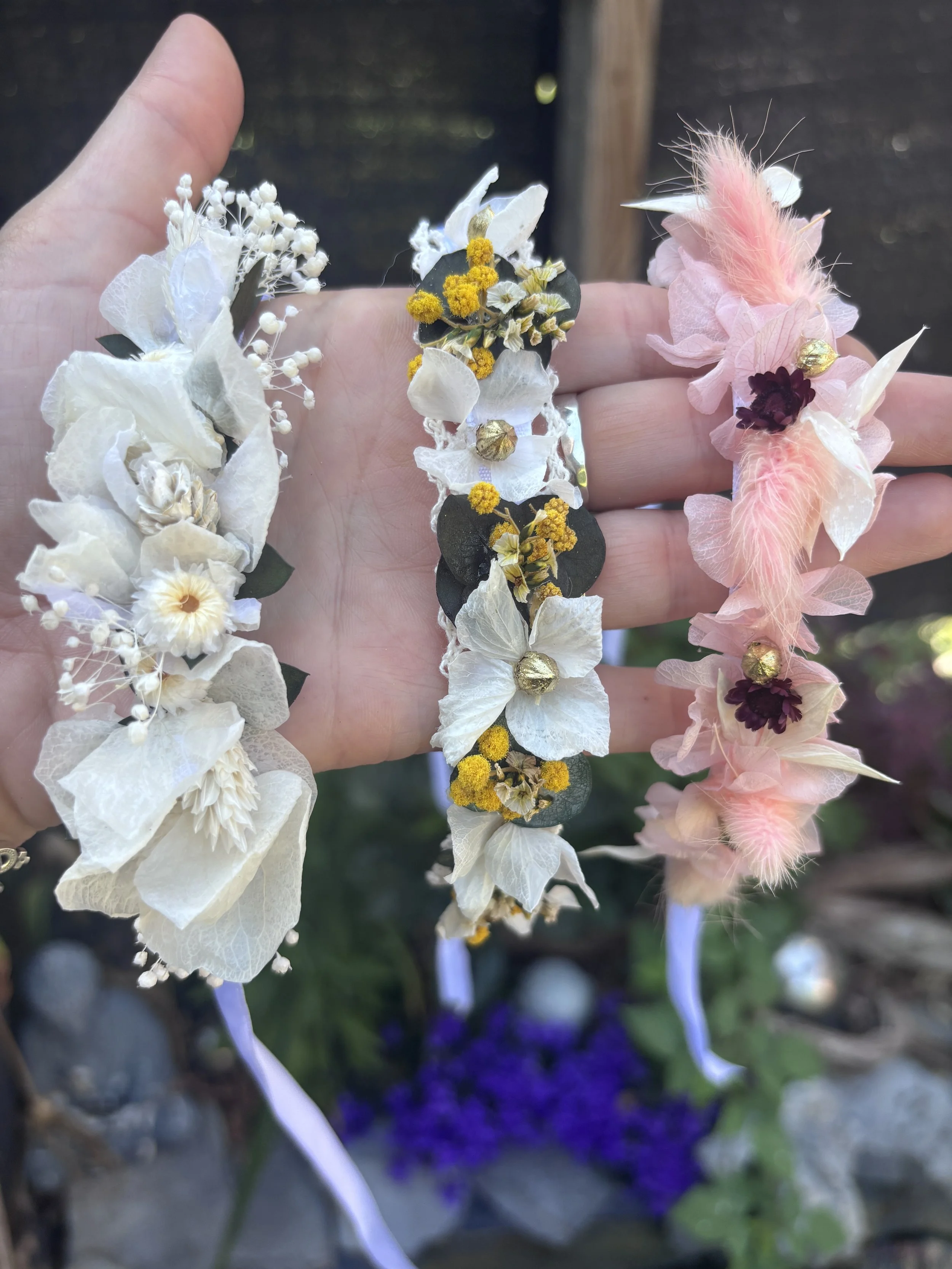 Bracelets de fleurs avec fleurs blanches, jaunes et roses, ornés de petits détails dorés et de plumes, porté sur une main.