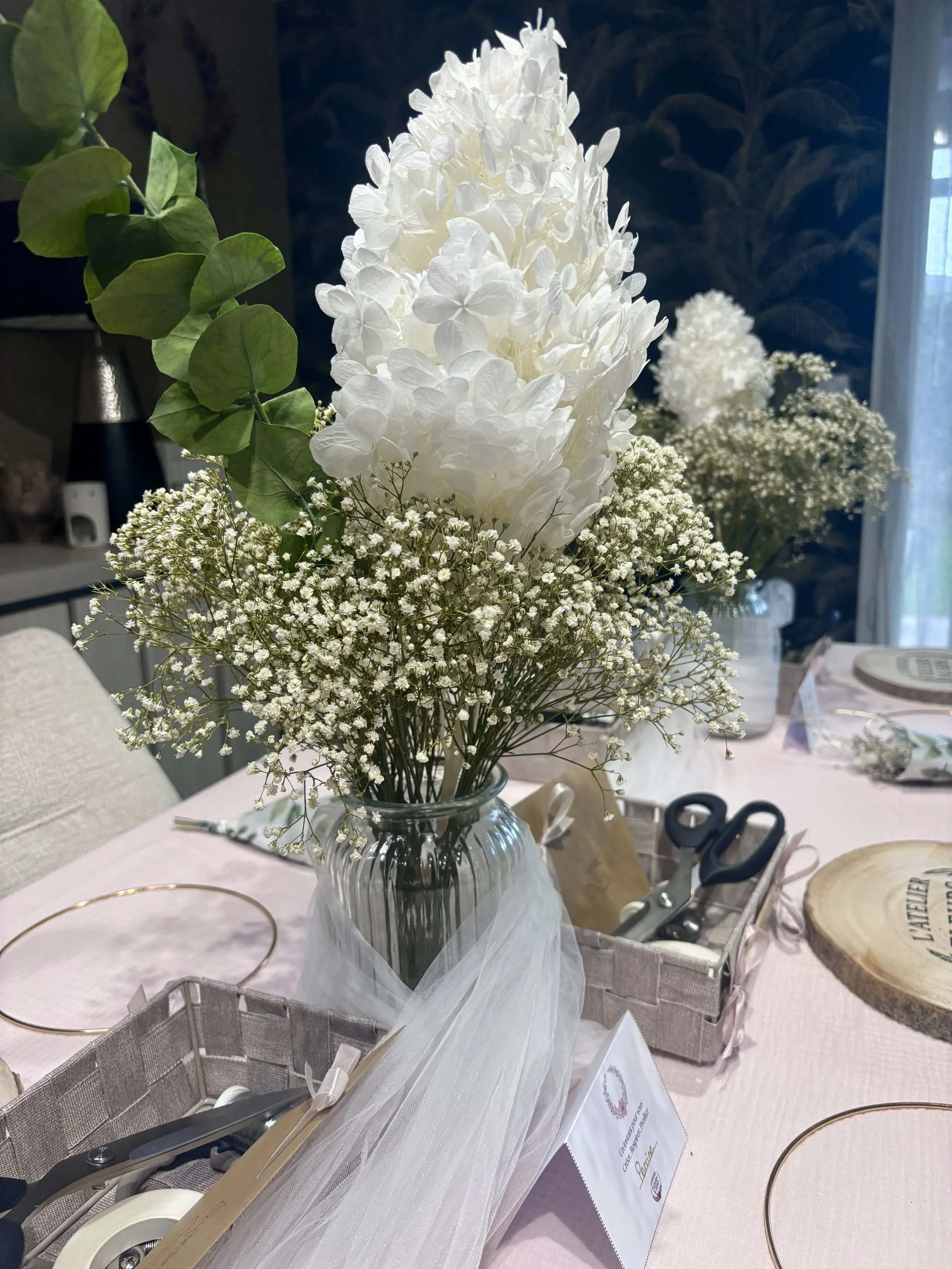 Centre de table floral avec des fleurs blanches, comprenant une grande tige de fleurs de hortensia blanches, du gypsophile et des feuilles vertes, dans un vase en verre. Table décorée avec un tissu blanc, des outils de bricolage, une boîte en tissu et un porte-assiette en métal.