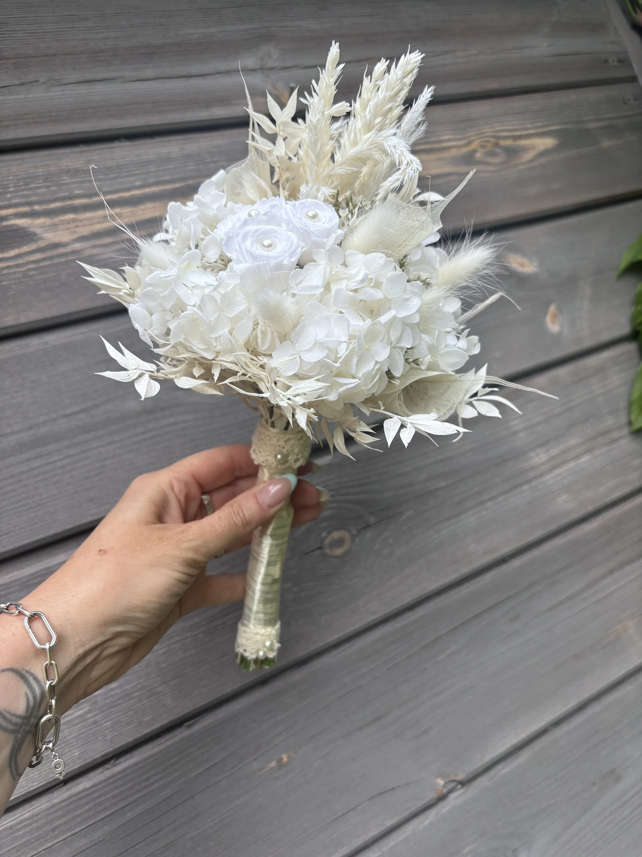 Bouquet de mariée en fleurs séchées et stabilisées blanches, réalisé artisanalement dans un esprit bohème chic et romantique. Cette création florale associe hortensias stabilisés, roses éternelles ornées de perles, arums délicats, feuillages blanchis