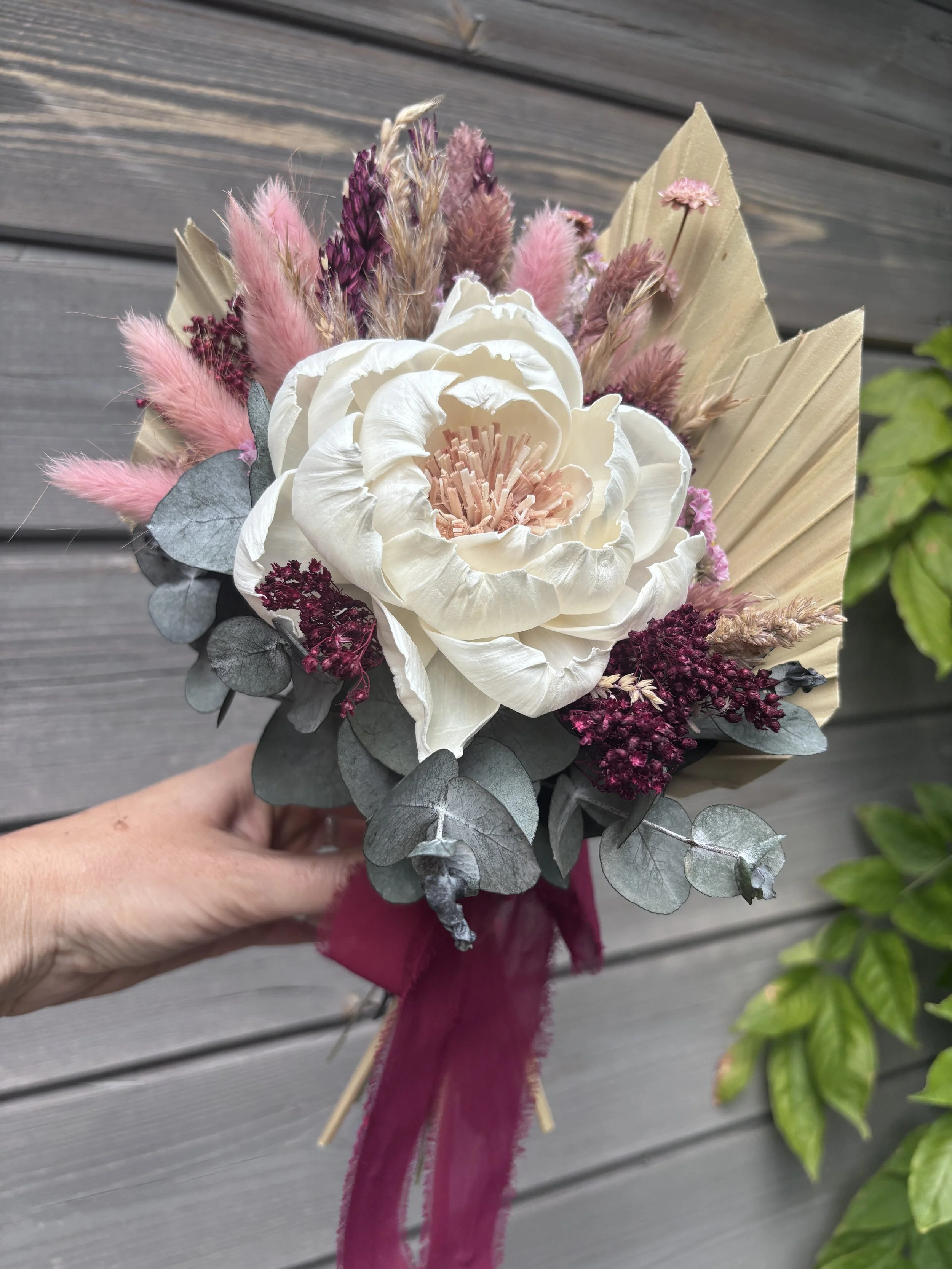 Bouquet de mariée en fleurs séchées et stabilisées, composé d’une grande pivoine ivoire en bois de Sola, d’eucalyptus, de lagurus rose poudré, de fleurs bordeaux et de palmes naturelles. Un bouquet romantique et durable, idéal pour un mariage bohème 