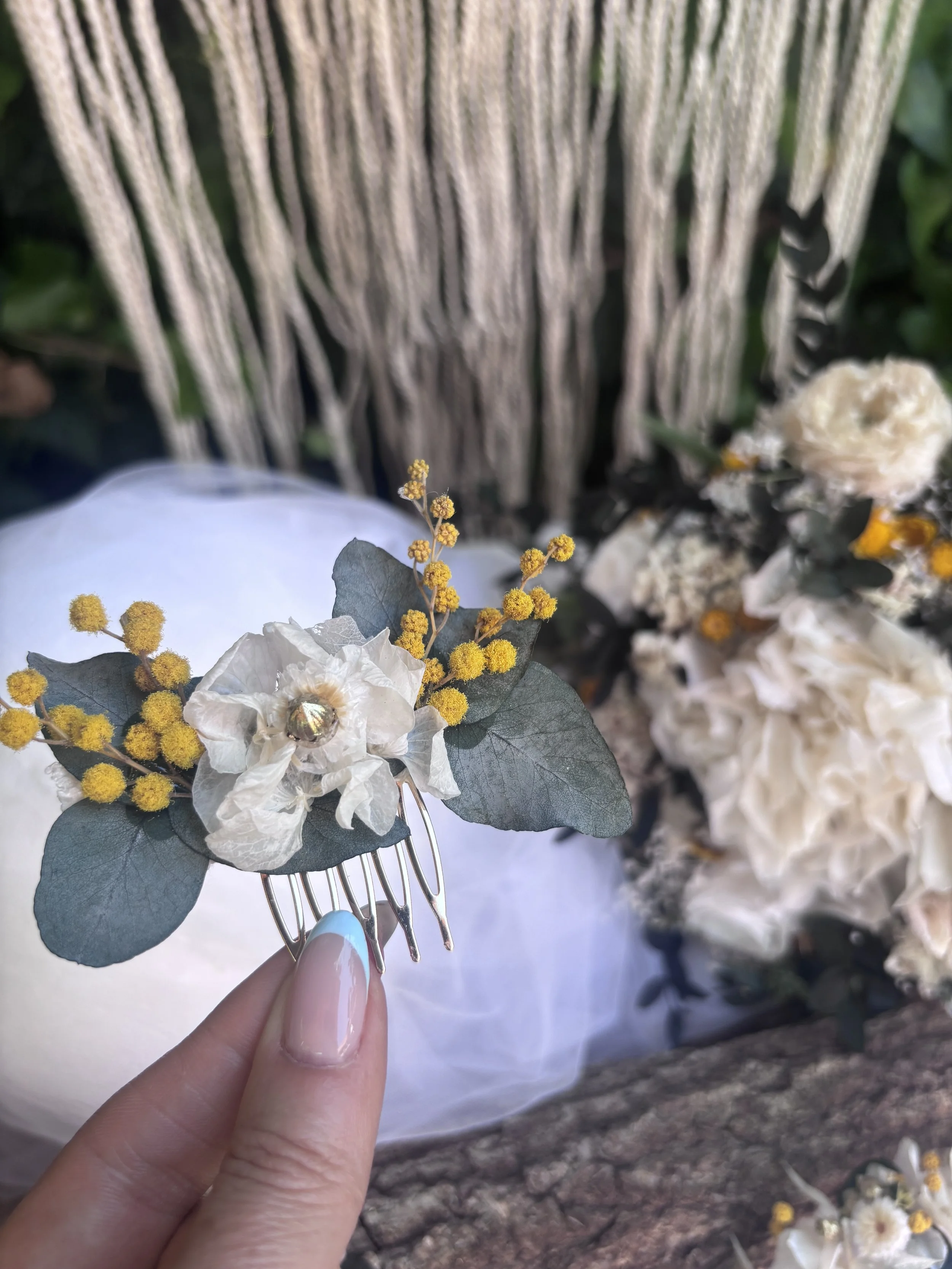 Main tenant une fourchette avec une décoration florale composée de fleurs blanches, de feuilles grisées et de petites fleurs jaunes, devant une toile de fond arborée.