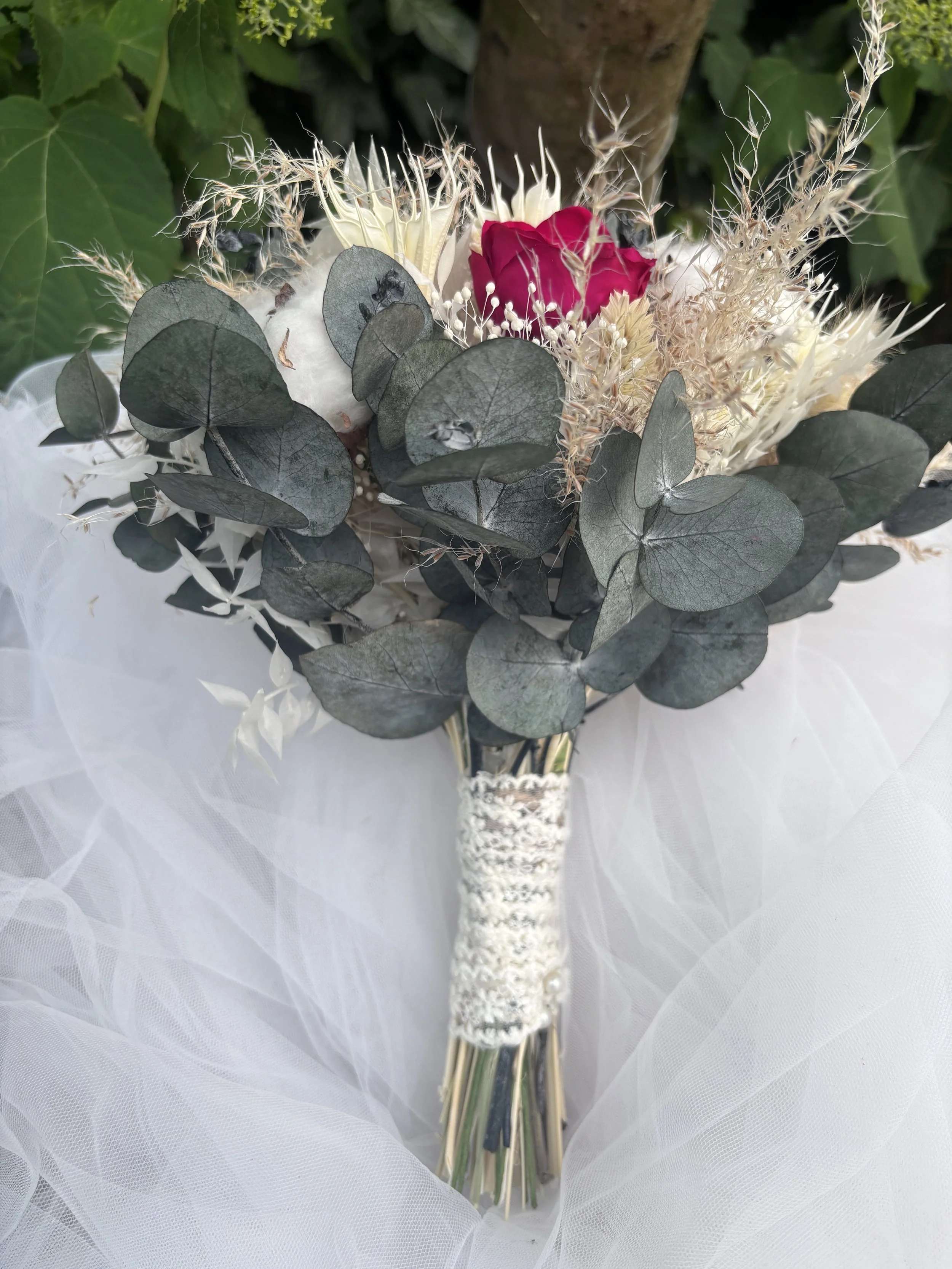 Bouquet de mariée bohème aux nuances bordeaux profond, ivoire et beige naturel, réalisé à partir de fleurs séchées et stabilisées. Cette création florale associe roses stabilisées bordeaux, coton, eucalyptus, graminées aériennes et fleurs texturées p