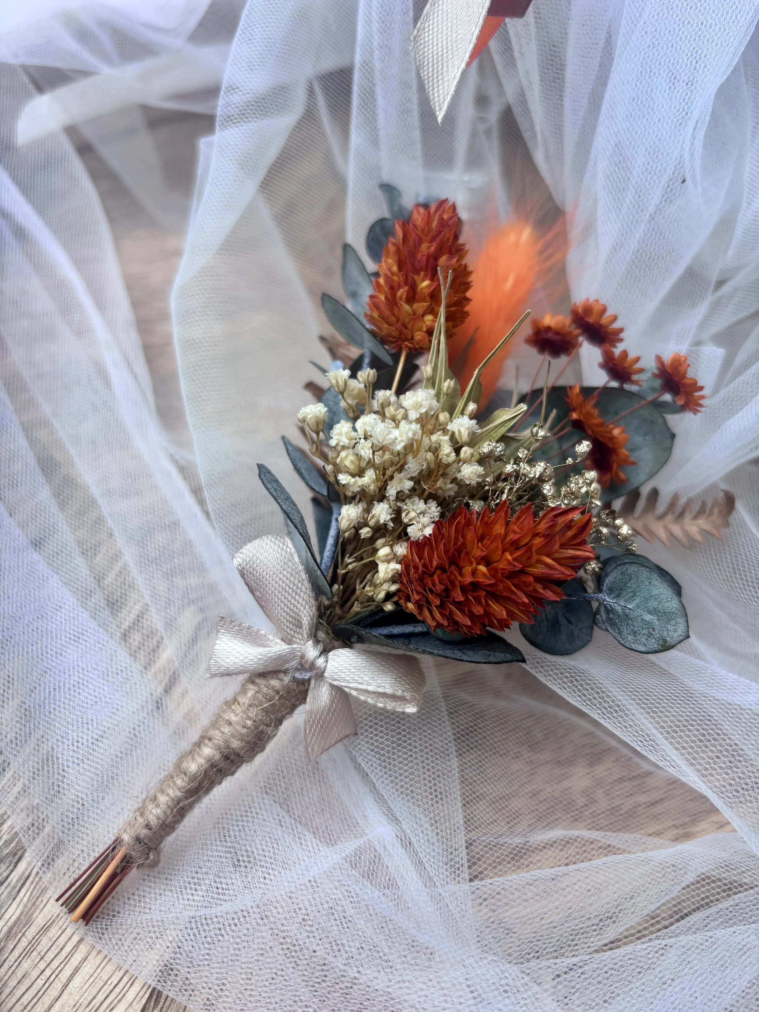 Boutonnière de marié en fleurs séchées et stabilisées, aux tons terracotta, orange, ivoire et eucalyptus, avec une touche dorée, idéale pour un mariage bohème.