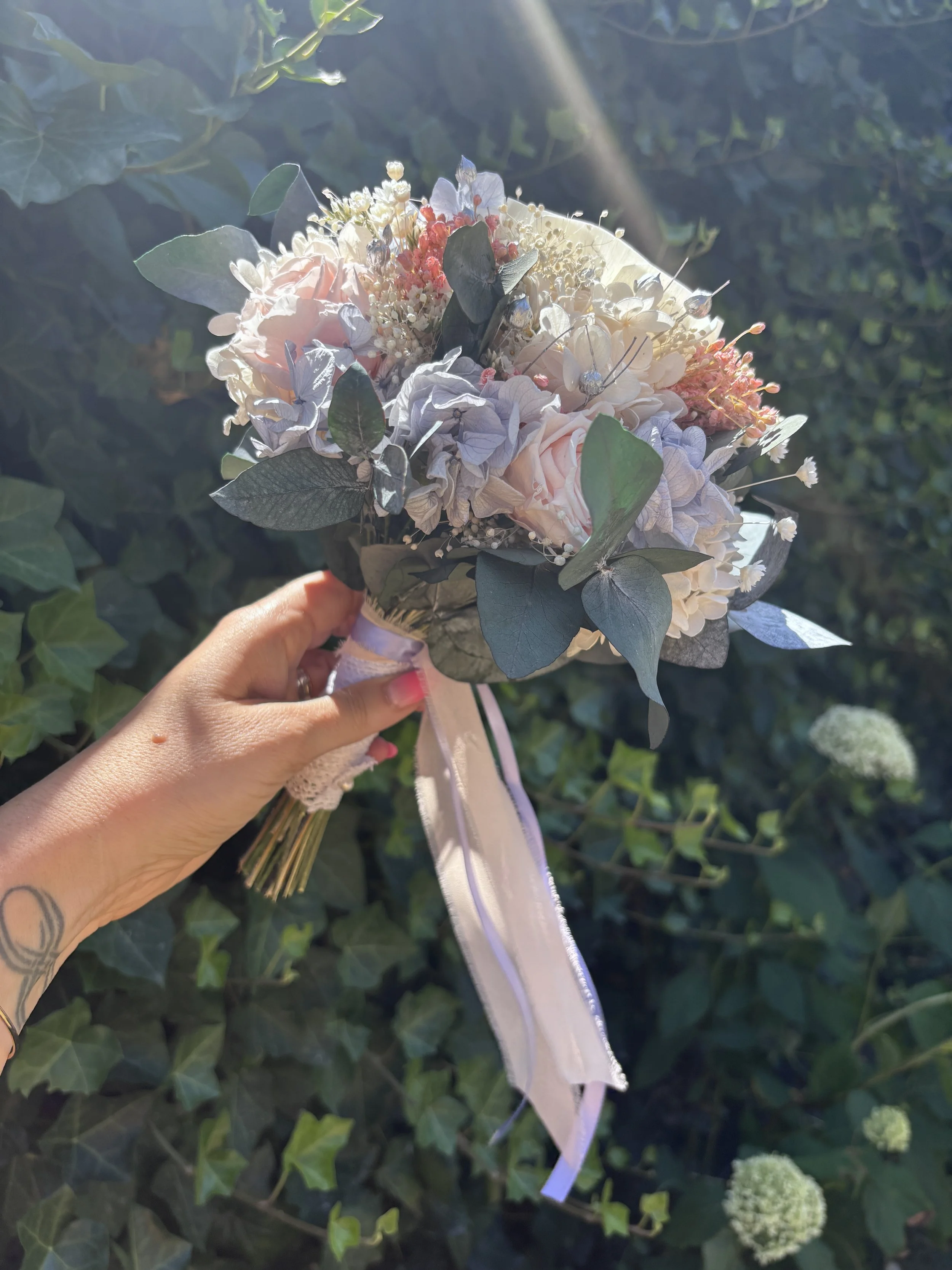 Bouquet de mariée en fleurs séchées et stabilisées aux teintes bleu pastel, rose poudré et ivoire, composé d’un camélia blanc en bois de Sola, d’hortensias stabilisés, de gypsophile, d’eucalyptus bleuté et de touches argentées. Une création romantiqu