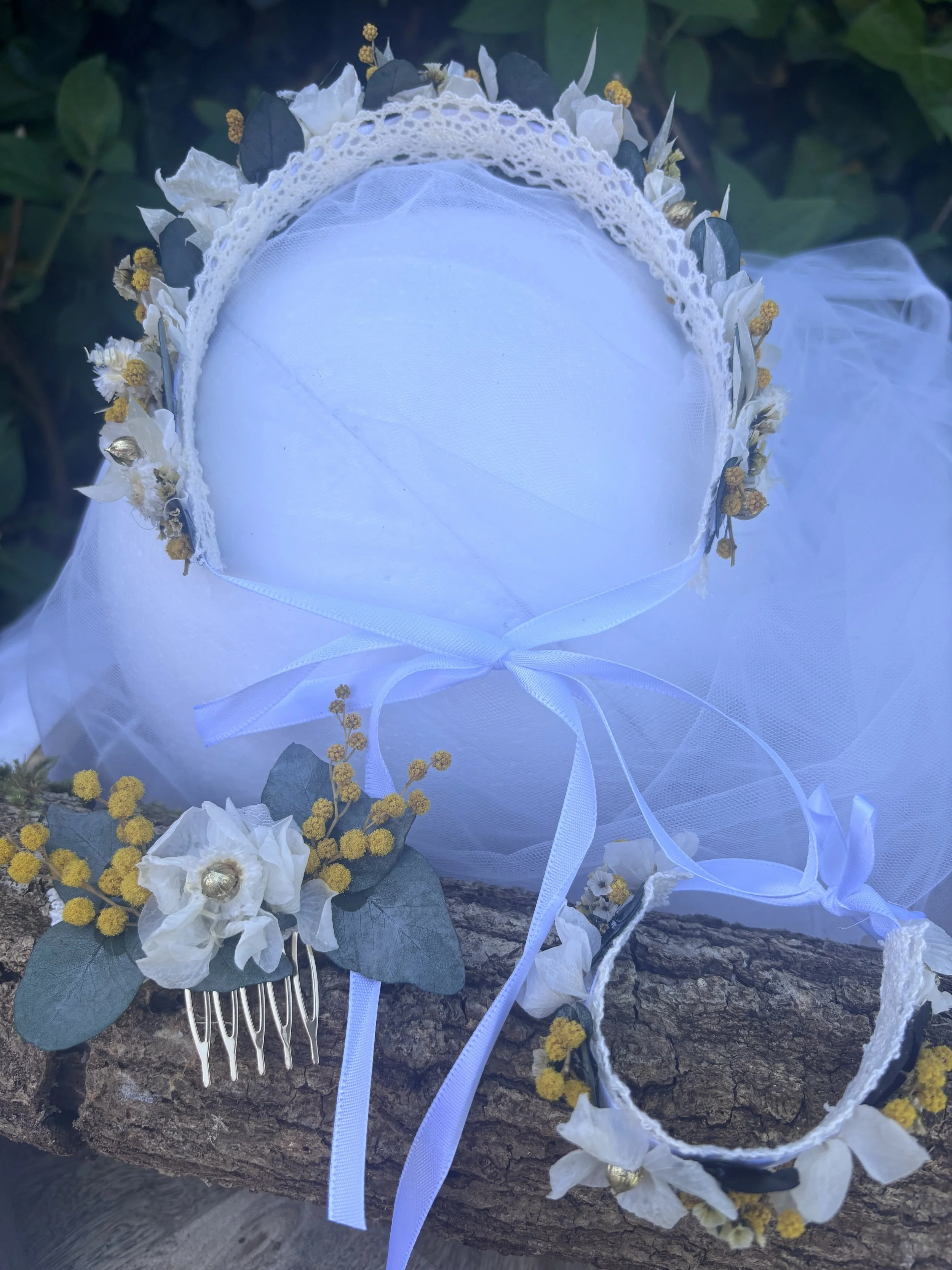 Une coiffure de mariée avec des fleurs blanches, des feuilles vertes, une fine dentelle, un voile blanc et une brosse à cheveux en arrière-plan.
