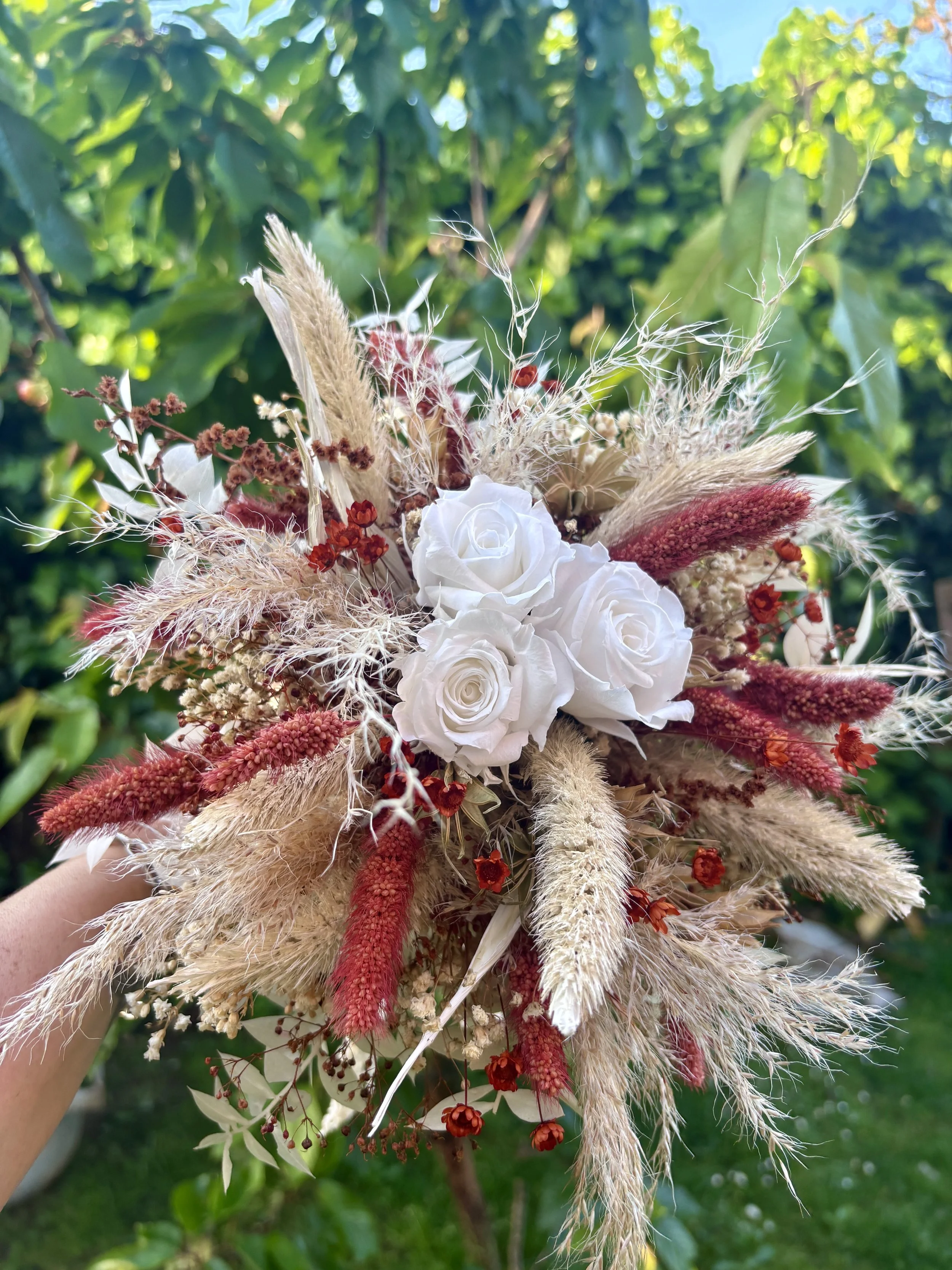 Bouquet de mariée bohème en fleurs séchées et stabilisées aux teintes terracotta, bordeaux, crème et blanc. Cette création florale artisanale associe roses éternelles blanches, pampas, setaria, graminées naturelles et délicates fleurs séchées pour of