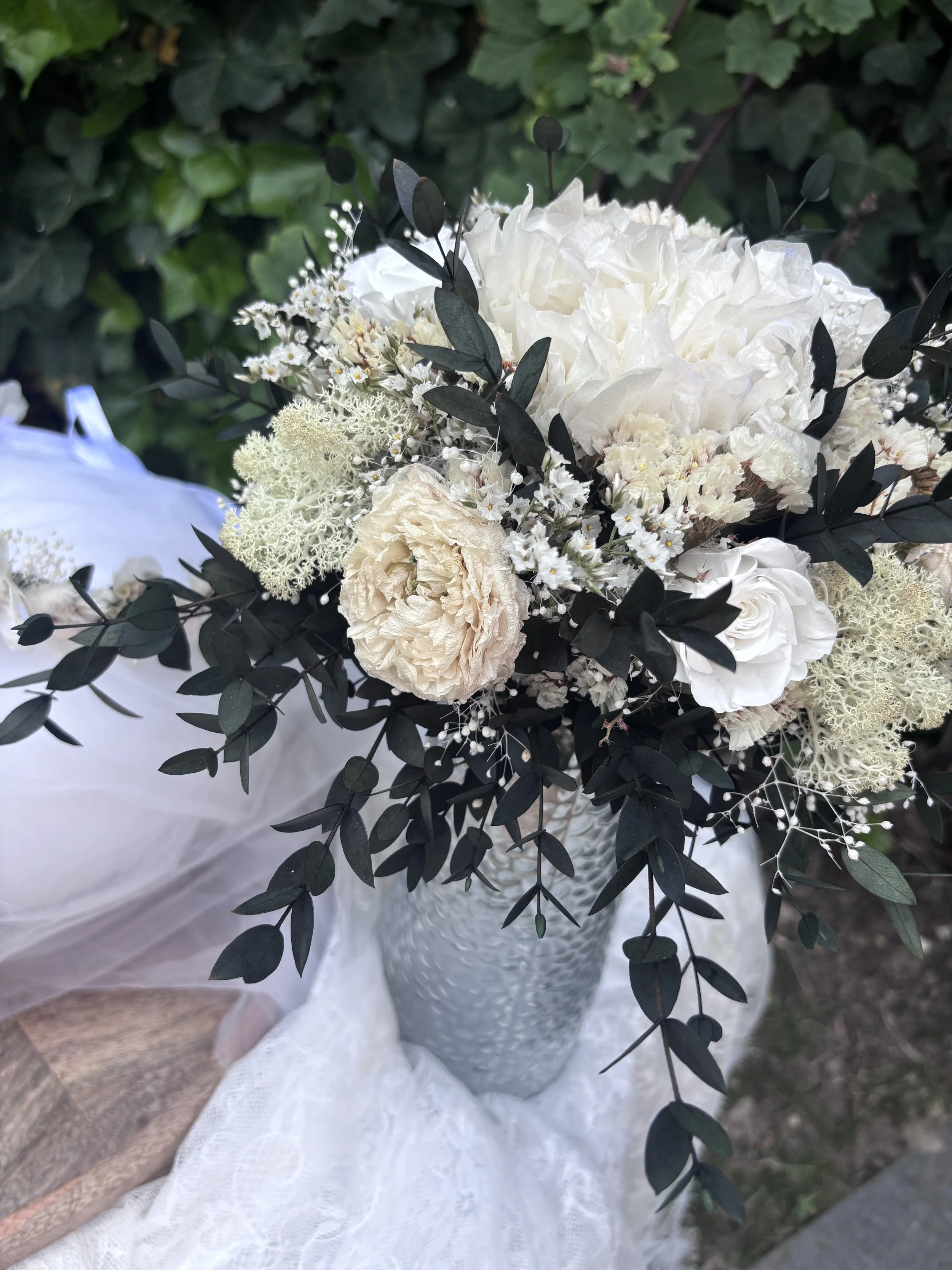 Bouquet de mariée bohème chic en fleurs séchées et stabilisées, composé de pivoines blanches, roses stabilisées, statice blanc, mousse végétale et feuillage d’eucalyptus noir. Cette création artisanale associe élégance, douceur et caractère pour un m