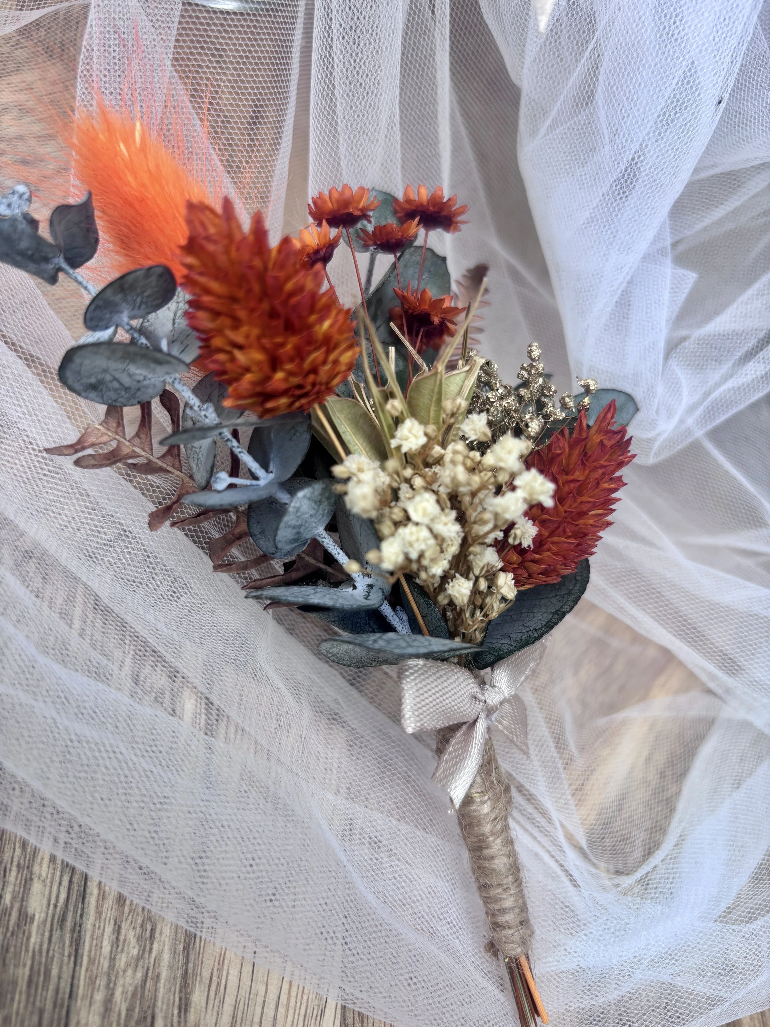 Boutonnière de marié en fleurs séchées et stabilisées, aux tons terracotta, orange, ivoire et eucalyptus, avec une touche dorée, idéale pour un mariage bohème.