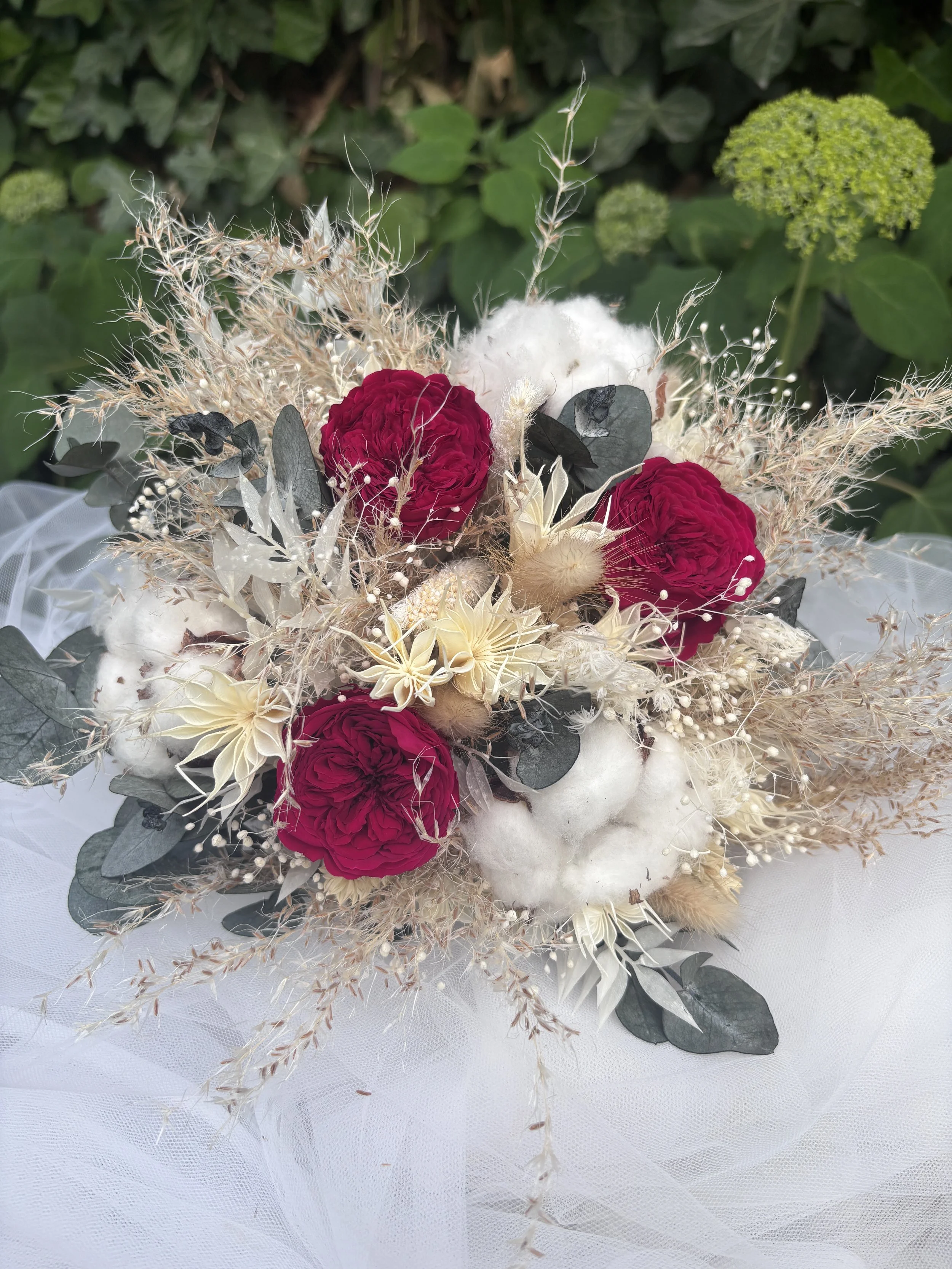 Bouquet de mariée bohème aux nuances bordeaux profond, ivoire et beige naturel, réalisé à partir de fleurs séchées et stabilisées. Cette création florale associe roses stabilisées bordeaux, coton, eucalyptus, graminées aériennes et fleurs texturées p