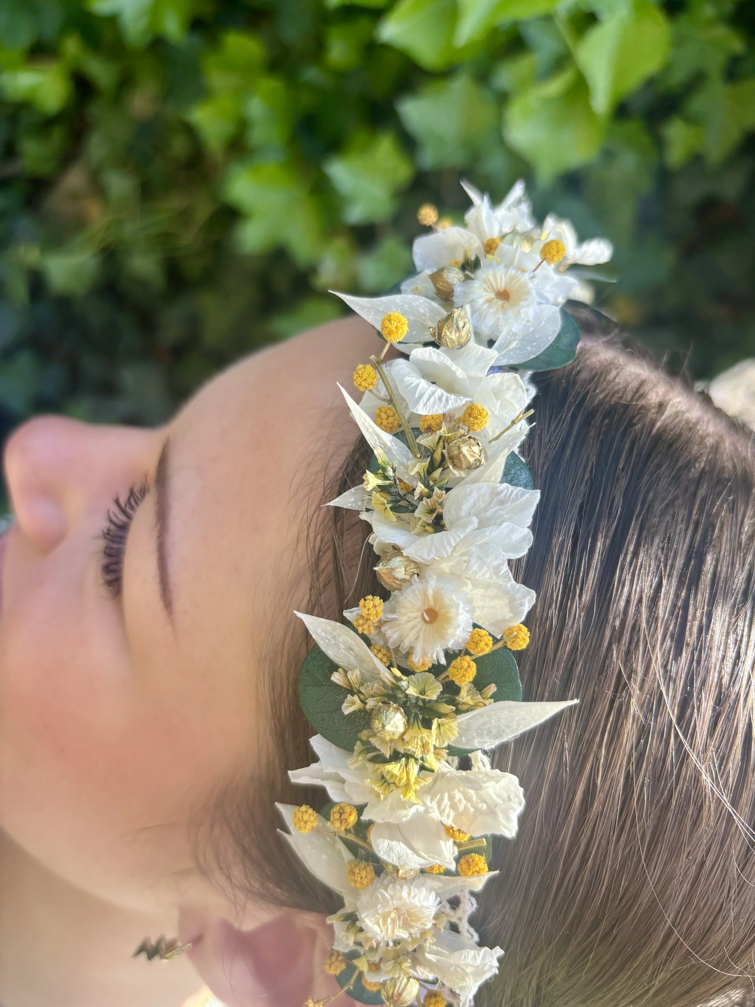 Vue latérale d'une femme avec une couronne de fleurs blanches et jaunes, cheveux longs et teints en gris clair, peau claire, dans un environnement naturel verdoyant.