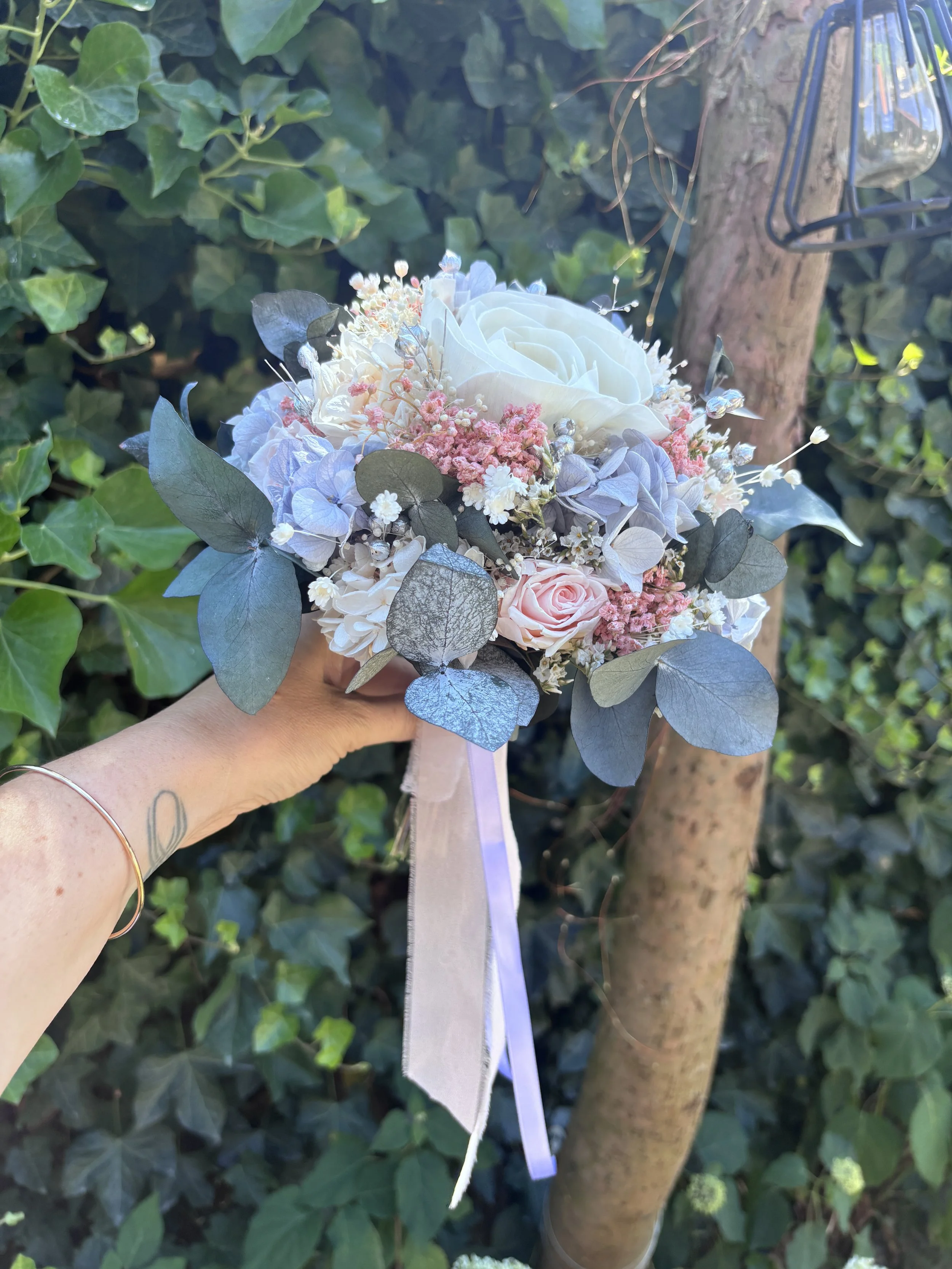 Bouquet de mariée en fleurs séchées et stabilisées aux teintes bleu pastel, rose poudré et ivoire, composé d’un camélia blanc en bois de Sola, d’hortensias stabilisés, de gypsophile, d’eucalyptus bleuté et de touches argentées. Une création romantiqu