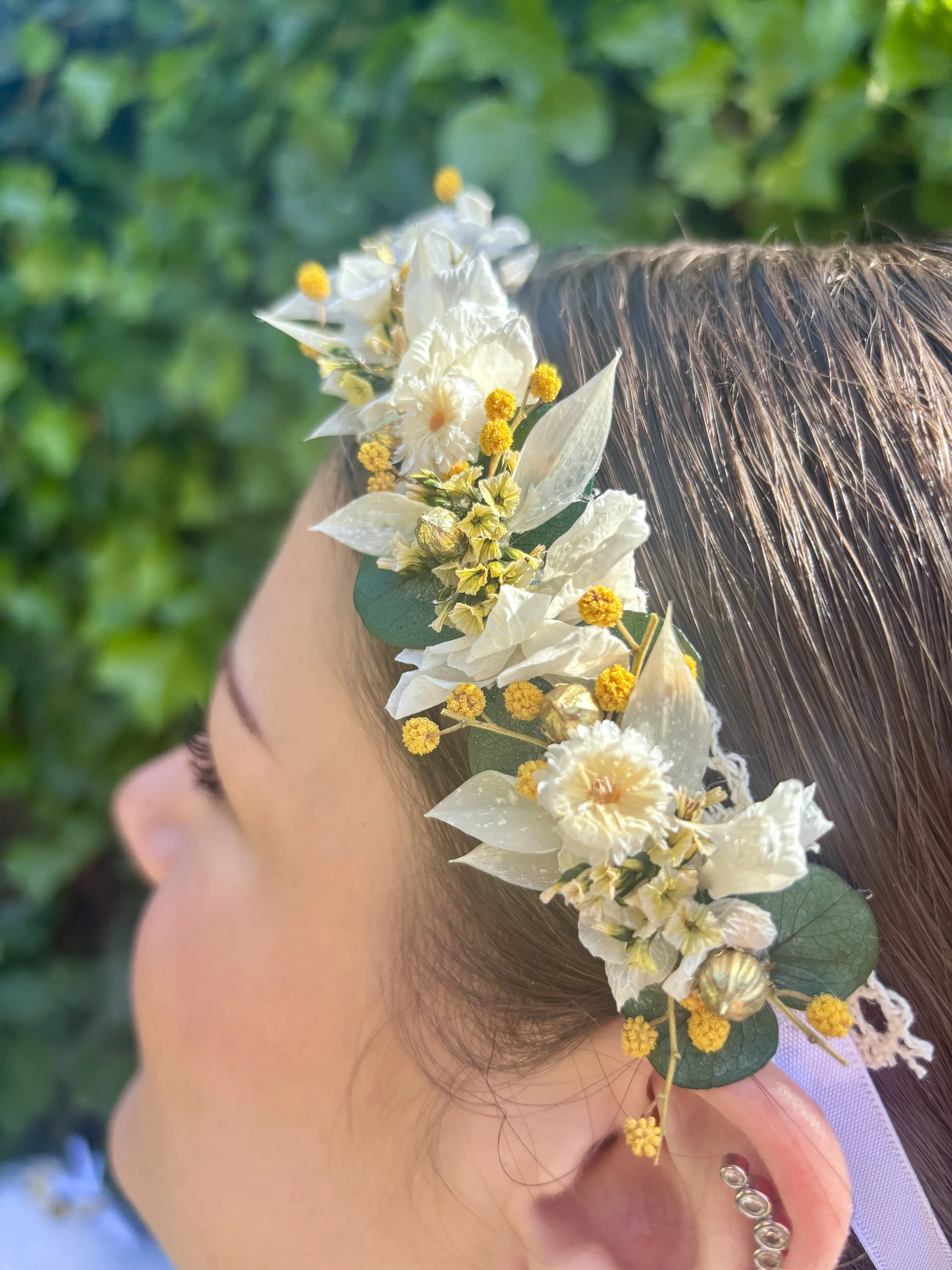 Une personne porte une couronne de fleurs blanches et jaunes autour de la tête, avec des feuilles vertes, dans un environnement naturel.