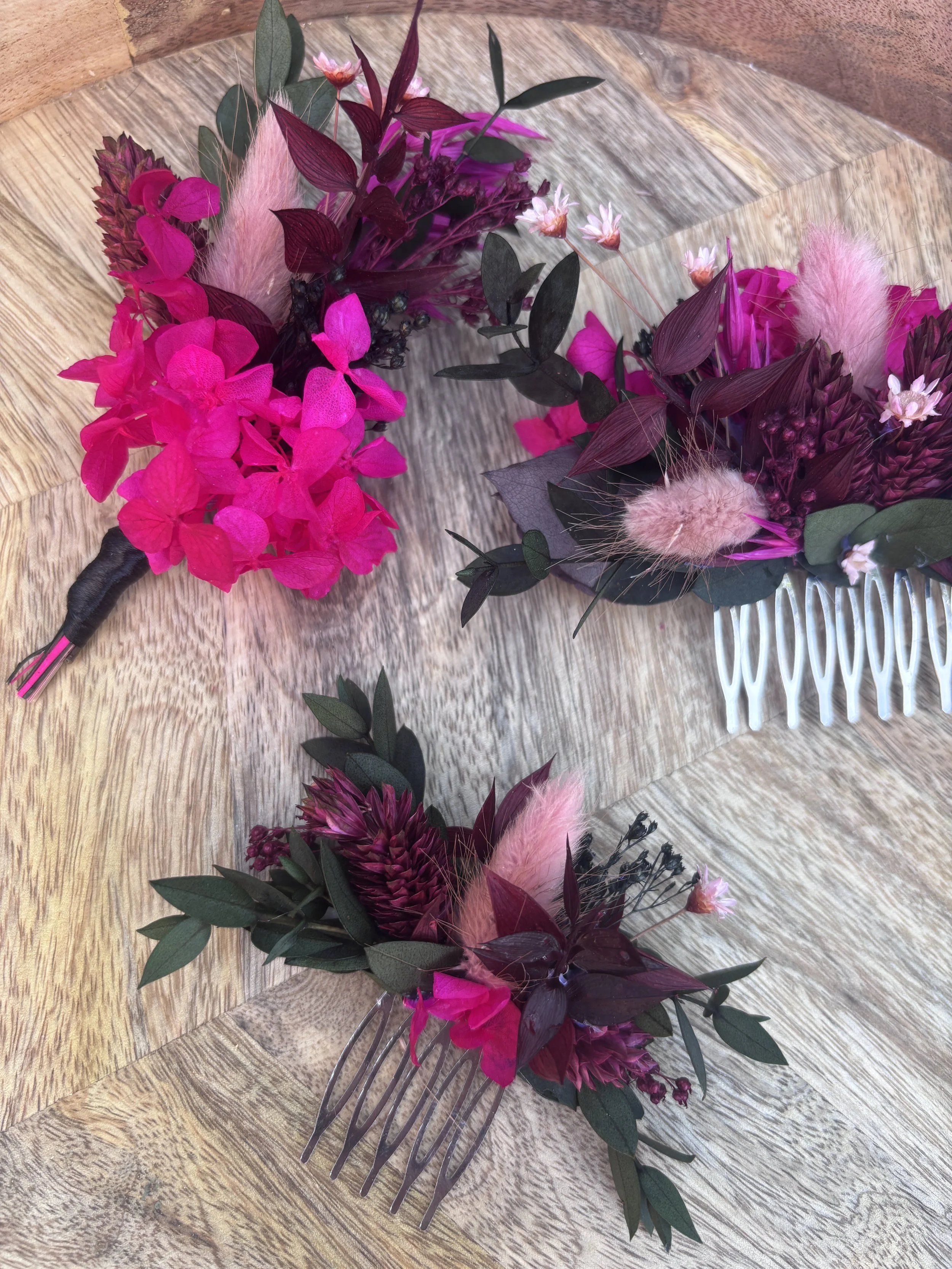 Couronne de fleurs avec des roses roses, des feuilles vertes, des plantes en plumes roses, et des fleurs blanches, posée sur une surface en bois.
