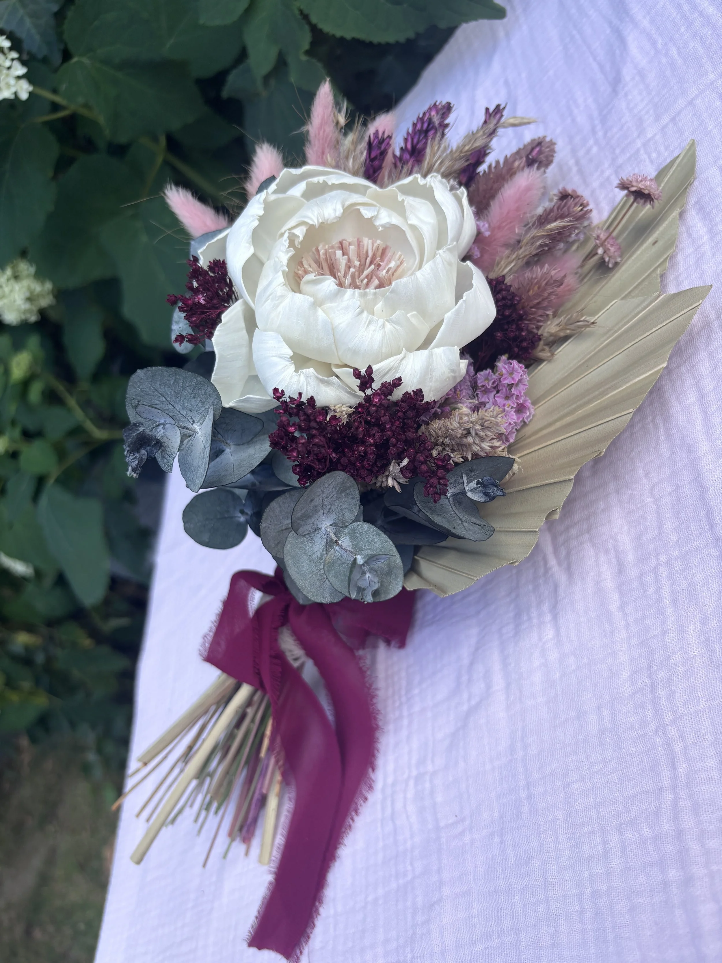 Bouquet de mariée en fleurs séchées et stabilisées, composé d’une grande pivoine ivoire en bois de Sola, d’eucalyptus, de lagurus rose poudré, de fleurs bordeaux et de palmes naturelles. Un bouquet romantique et durable, idéal pour un mariage bohème 