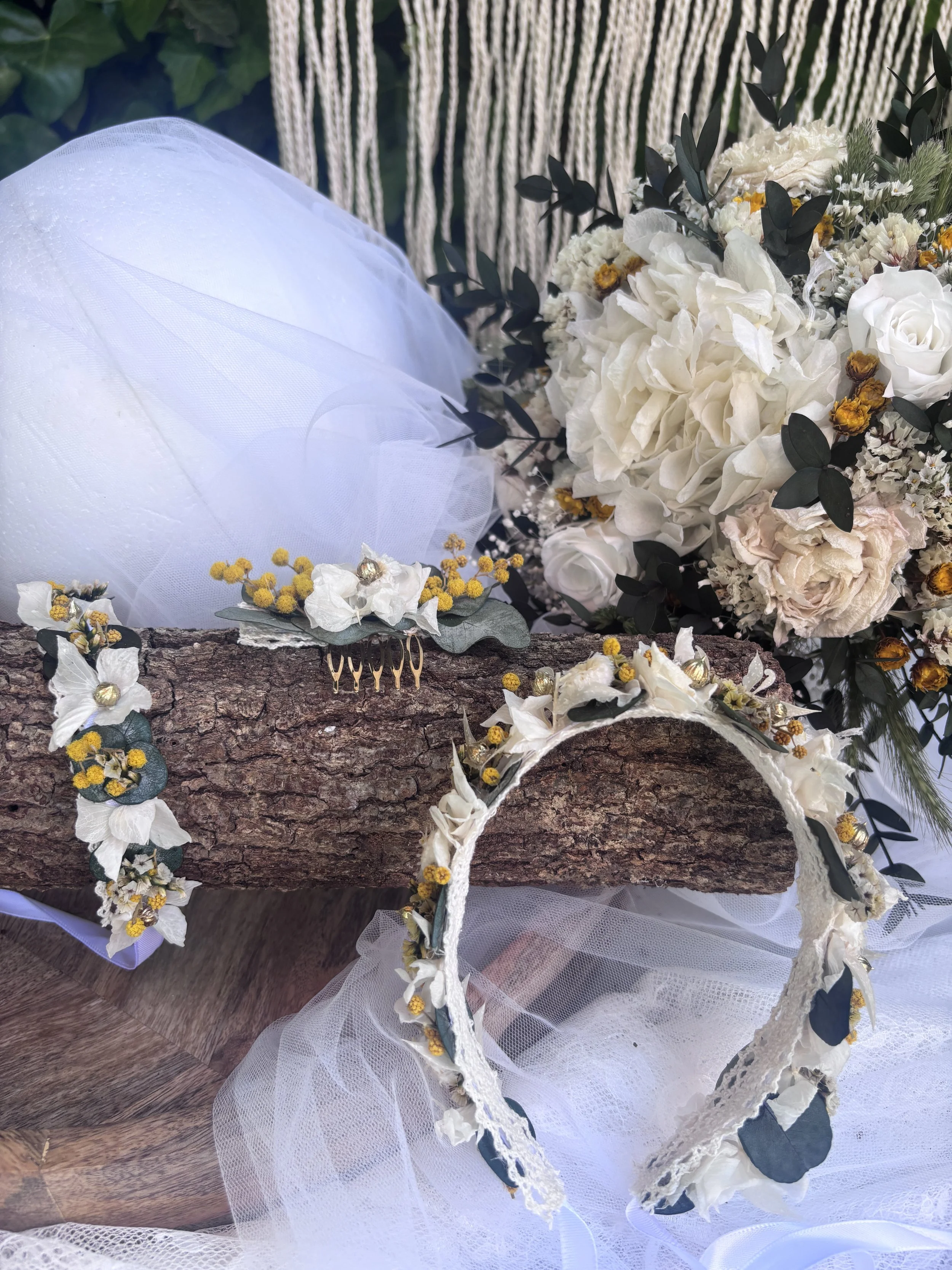 Une couronne de fleurs blanche avec des feuillages verts et jaunes, posée sur une branche de bois flotté, accompagnée d'une étole en tulle blanc, évoquant un décor de mariage.