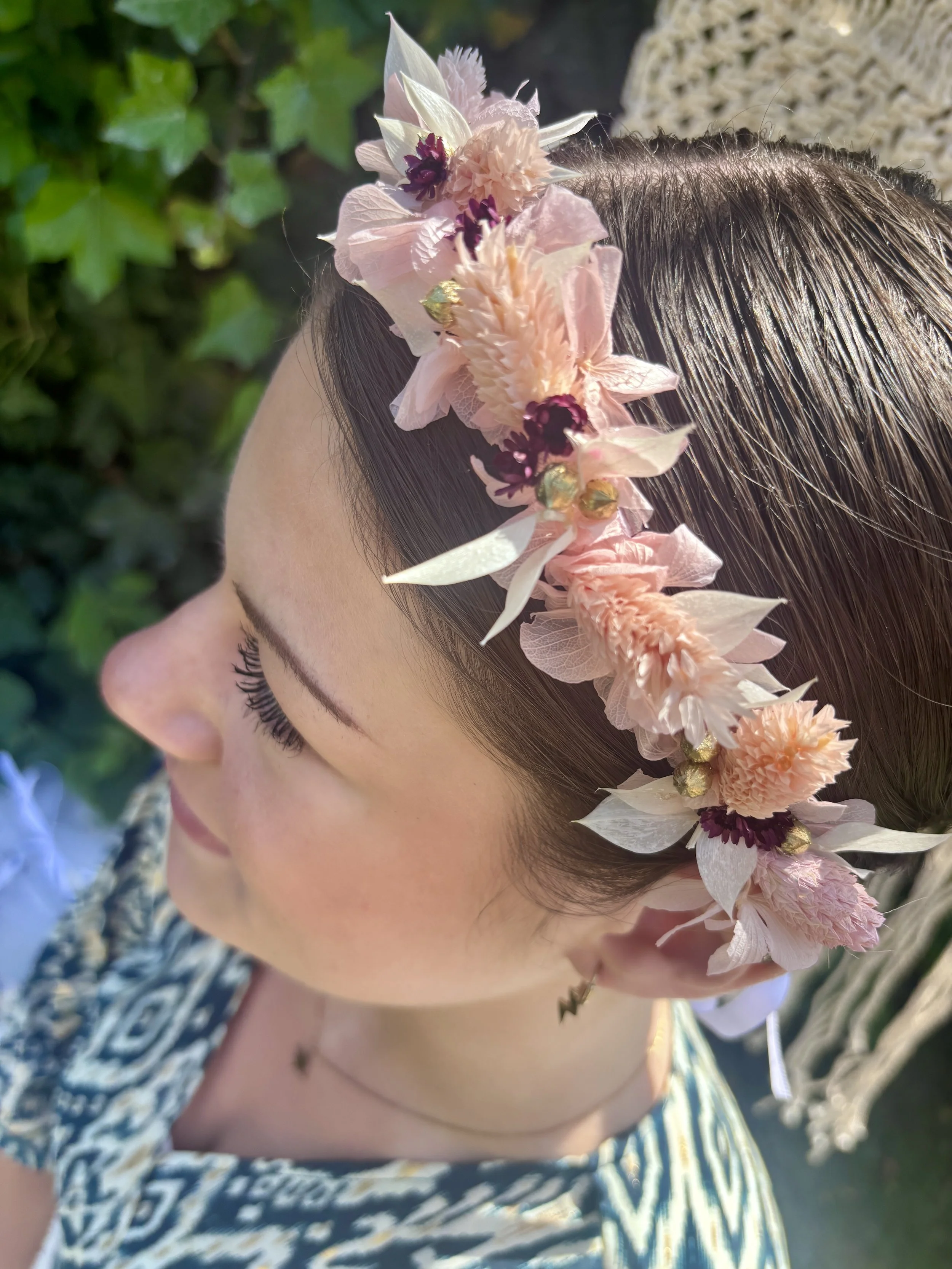 Une femme portant une couronne de fleurs dans ses cheveux, avec des fleurs roses, blanches et violettes, et un fond de verdure.