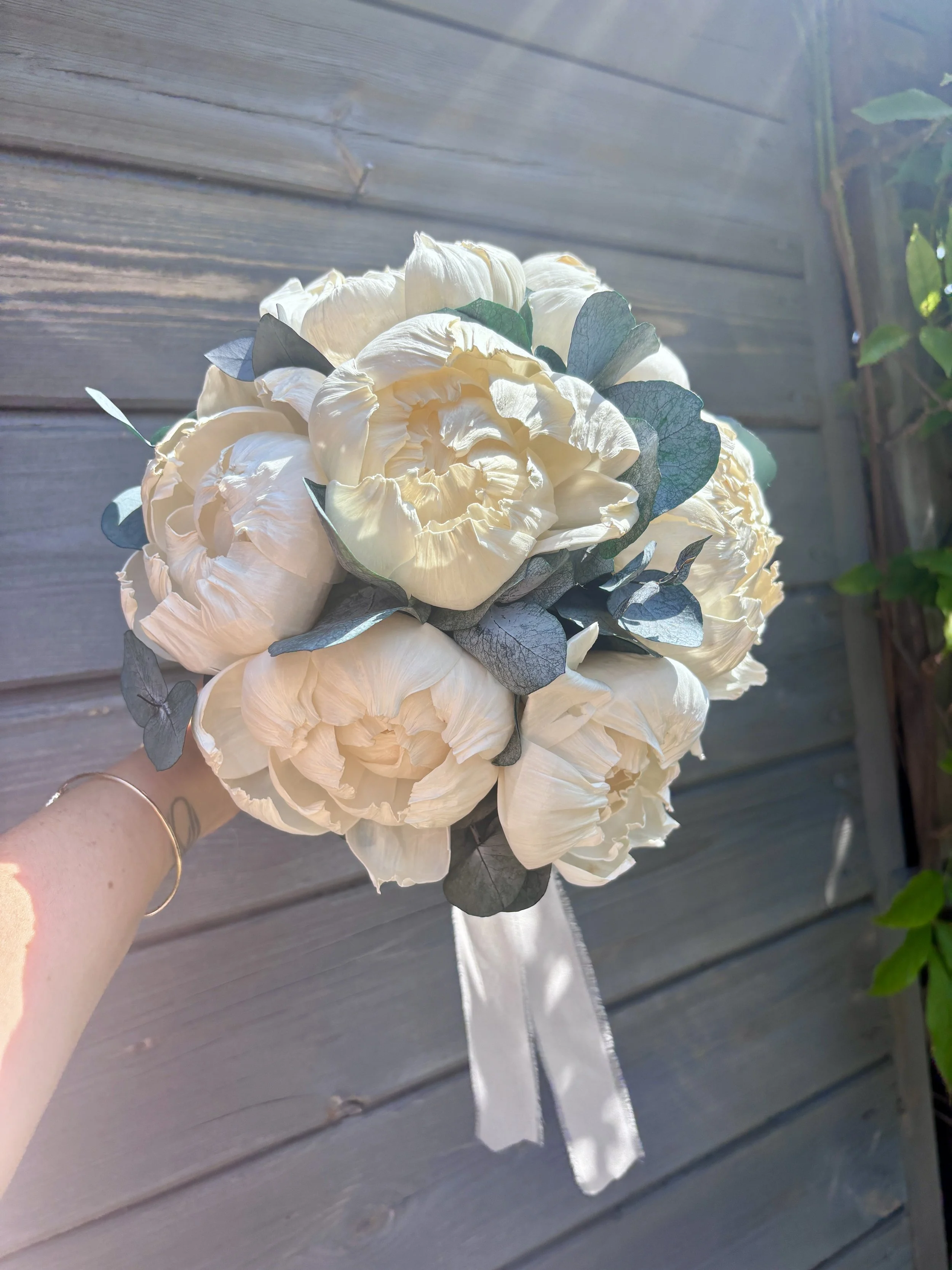 Bouquet de mariée élégant et intemporel composé de grandes pivoines ivoire en bois de Sola, associées à un feuillage d’eucalyptus. Une création florale durable, réalisée artisanalement et personnalisable selon les couleurs du mariage par L’Atelier Fl