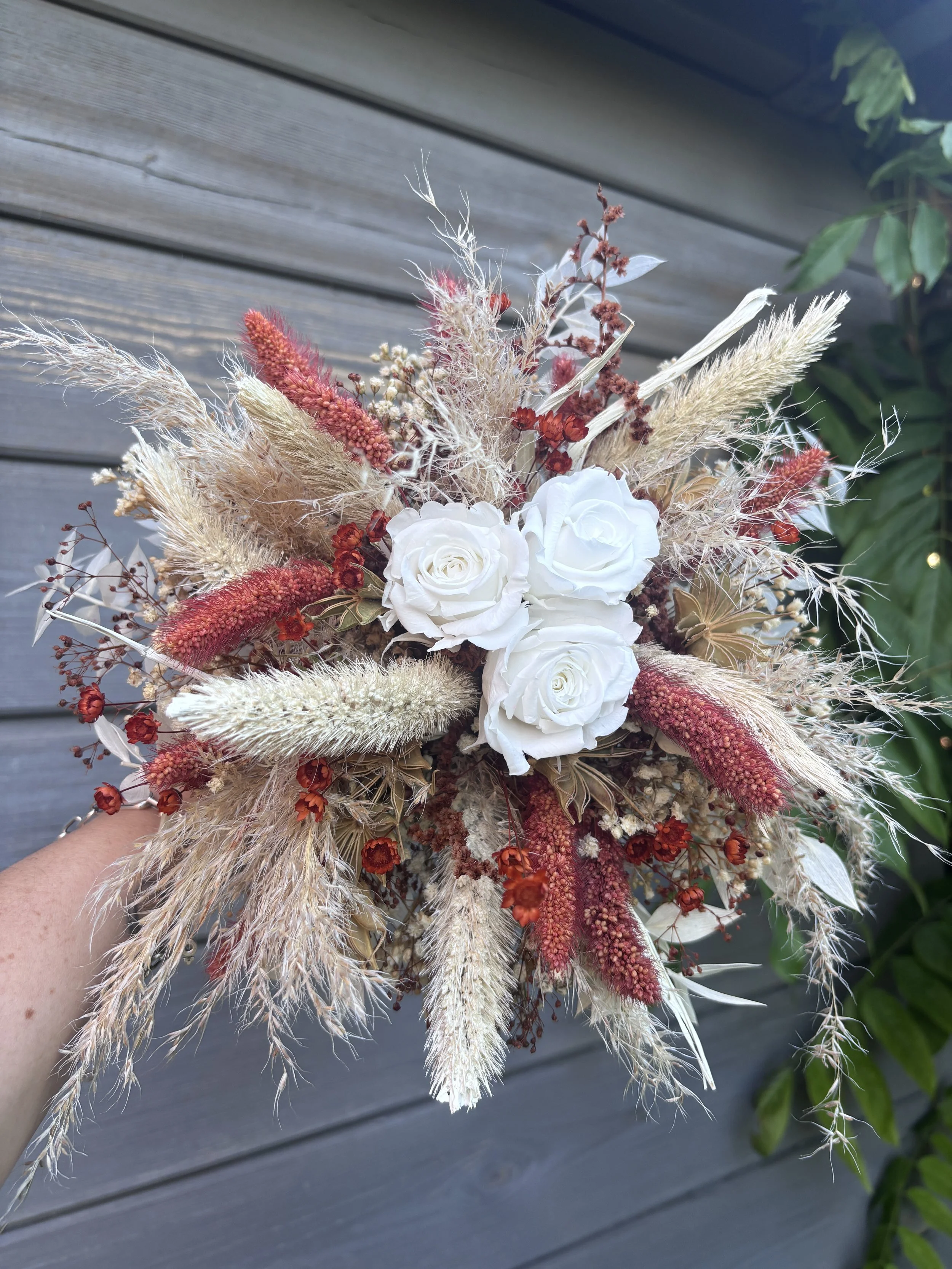 Bouquet de mariée bohème en fleurs séchées et stabilisées aux teintes terracotta, bordeaux, crème et blanc. Cette création florale artisanale associe roses éternelles blanches, pampas, setaria, graminées naturelles et délicates fleurs séchées pour of